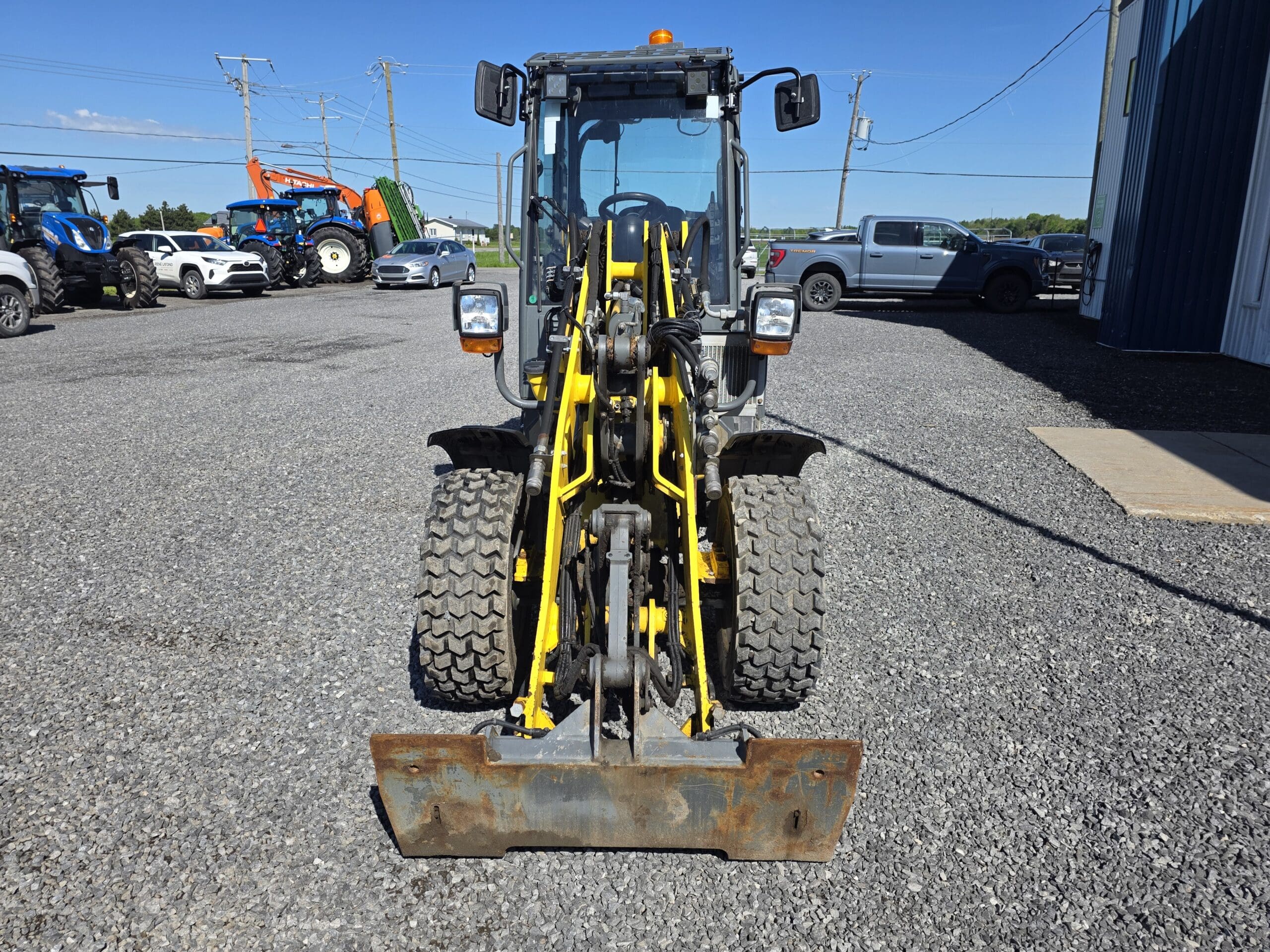 Vue de face d'une chargeuse de chantier jaune avec de gros pneus, garée sur un terrain en gravier avec des voitures, des camions et des tracteurs en arrière-plan sous un ciel bleu clair.