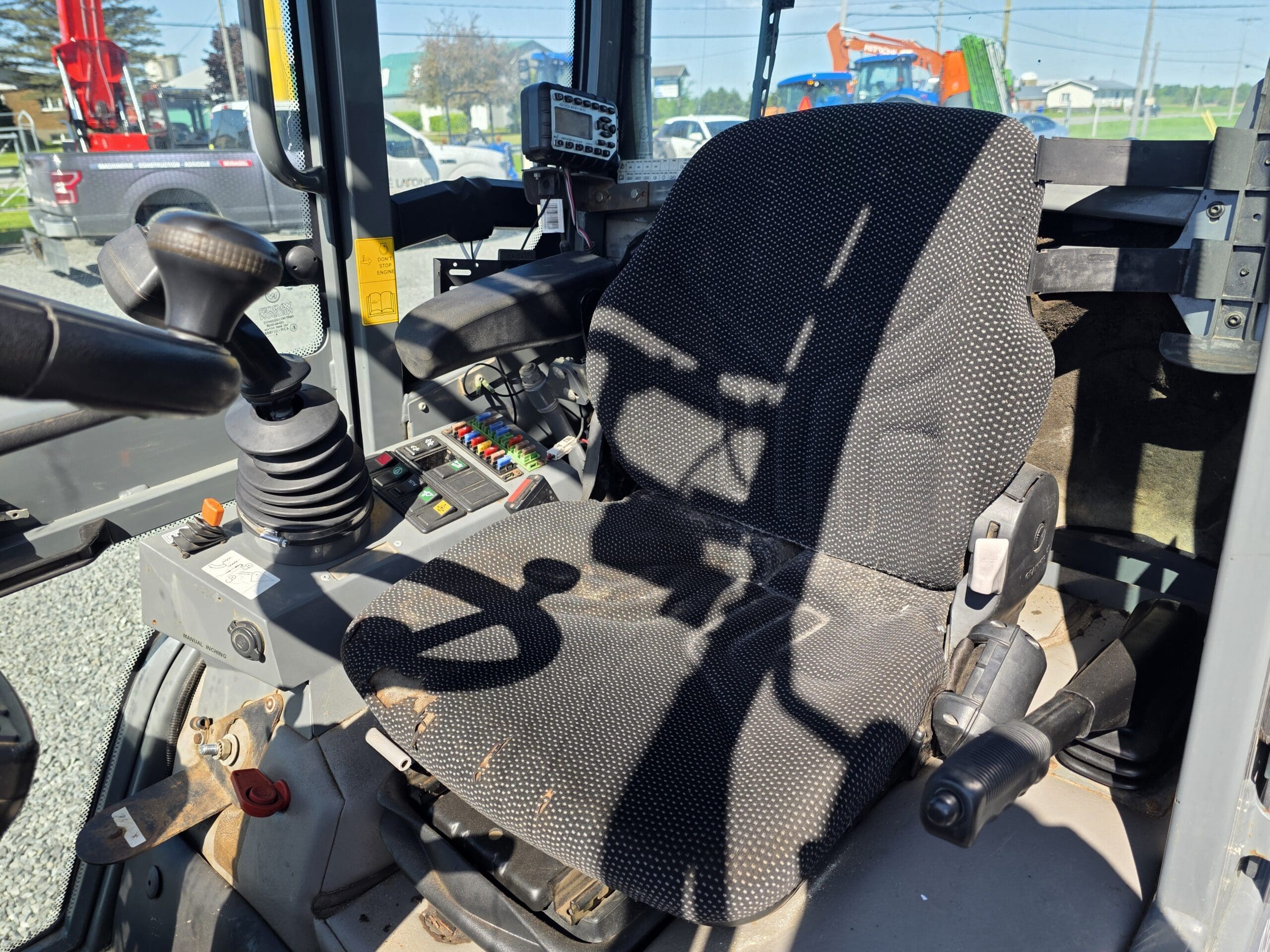 La photo montre le siège conducteur et le panneau de commande d'un engin de chantier, comme une excavatrice. Le siège est noir et entouré de leviers, de boutons et d'un joystick, tandis que la lumière du soleil projette des ombres sur la zone.
