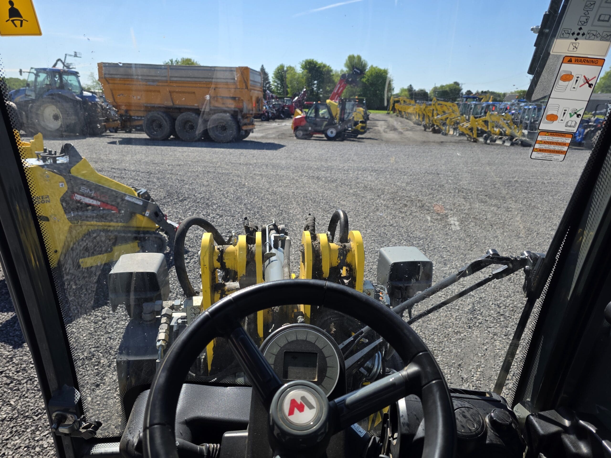 Vue de l'intérieur d'un véhicule de construction montrant le volant et les commandes, regardant les machines lourdes, les camions et l'équipement garés sur un terrain en gravier sous un ciel clair.