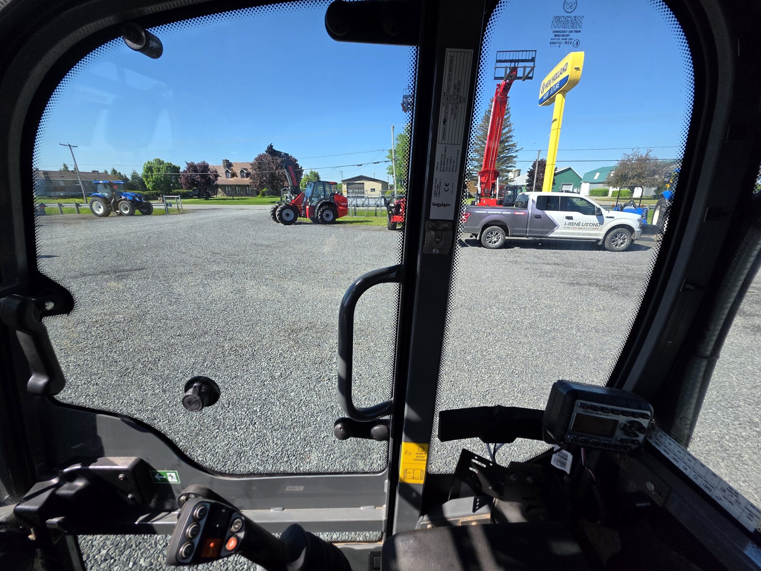 Vue depuis l'intérieur d'une cabine de véhicule donnant sur un terrain en gravier avec des tracteurs, un élévateur rouge et une camionnette blanche. Des bâtiments, des arbres et un panneau jaune sont visibles en arrière-plan sous un ciel bleu.