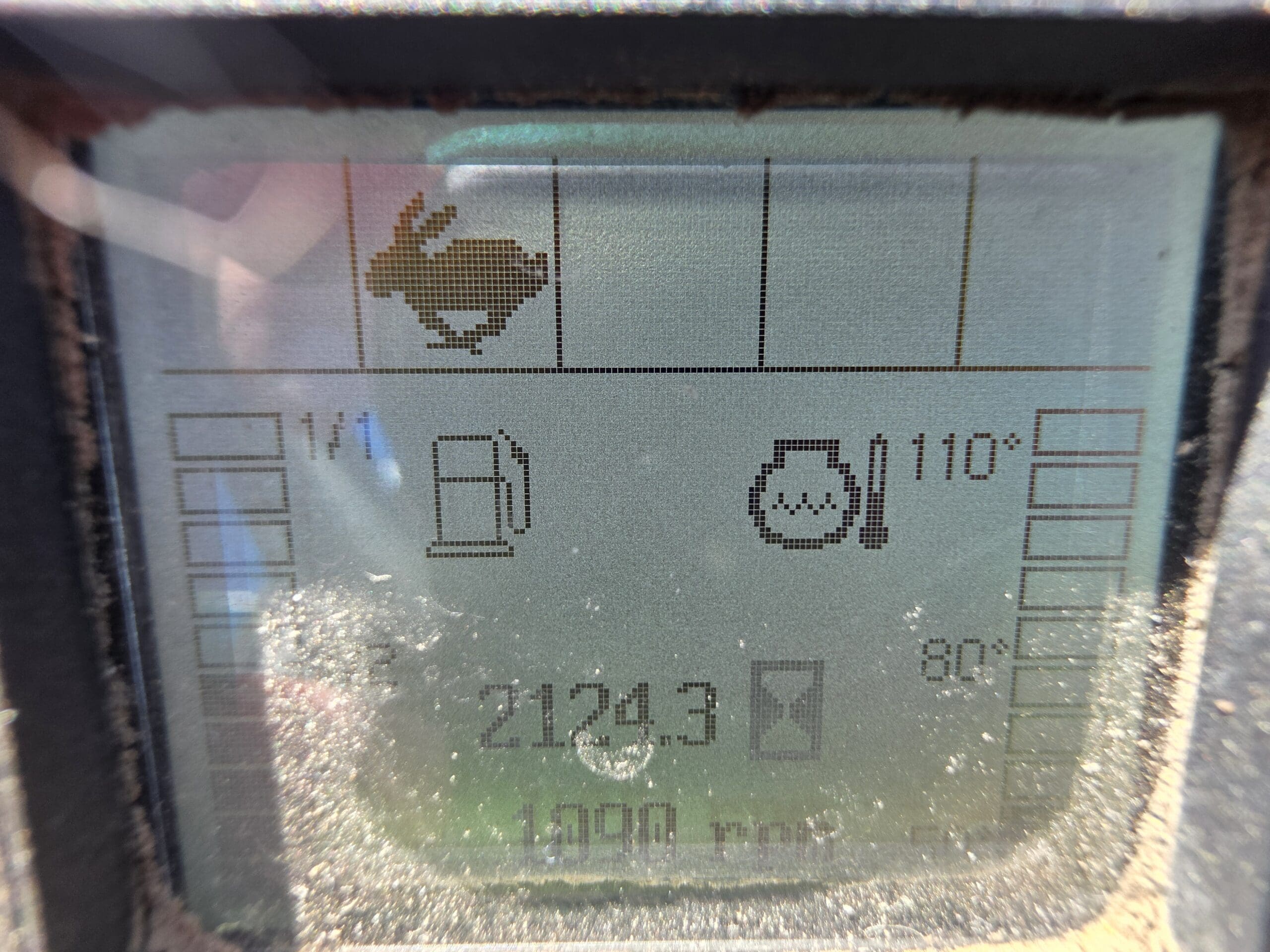 Gros plan d'un écran d'affichage numérique poussiéreux affichant une icône de lapin, une jauge de carburant, une lecture de température de 110° et diverses icônes et chiffres, dont « 2124,3 » près du bas.