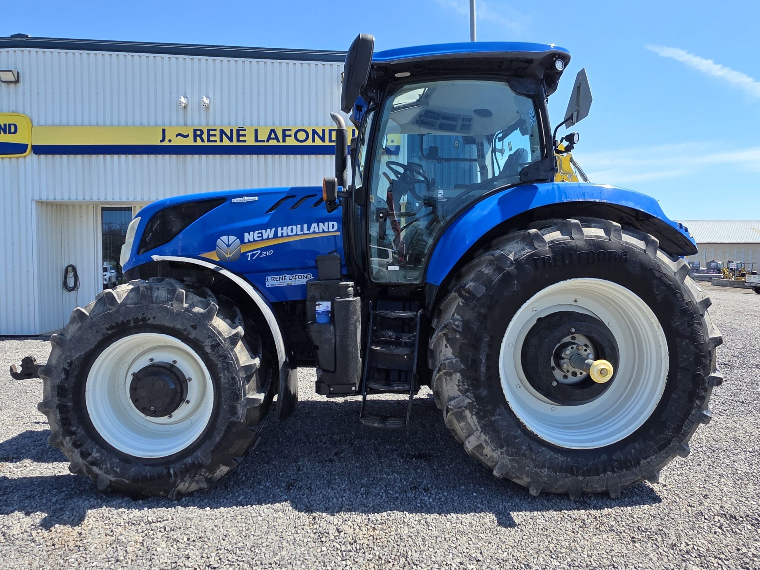 Un tracteur New Holland bleu est garé sur du gravier devant un bâtiment portant une pancarte « J. René Lafond ». Le tracteur est équipé de gros pneus noirs avec des jantes blanches et des détails de cabine visibles.