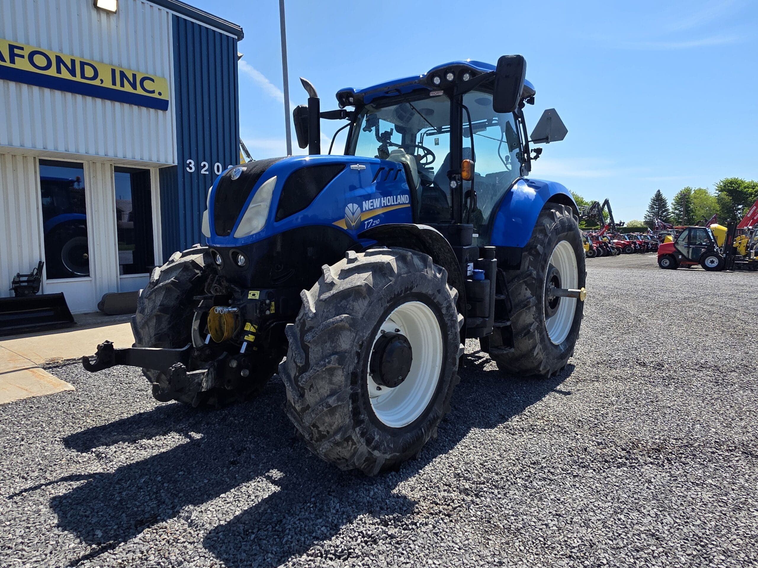 Un tracteur New Holland bleu est garé sur du gravier devant un bâtiment aux bardages blancs et bleus, sous un ciel dégagé. Le tracteur est légèrement orienté vers la gauche, et des équipements agricoles sont visibles en arrière-plan.
