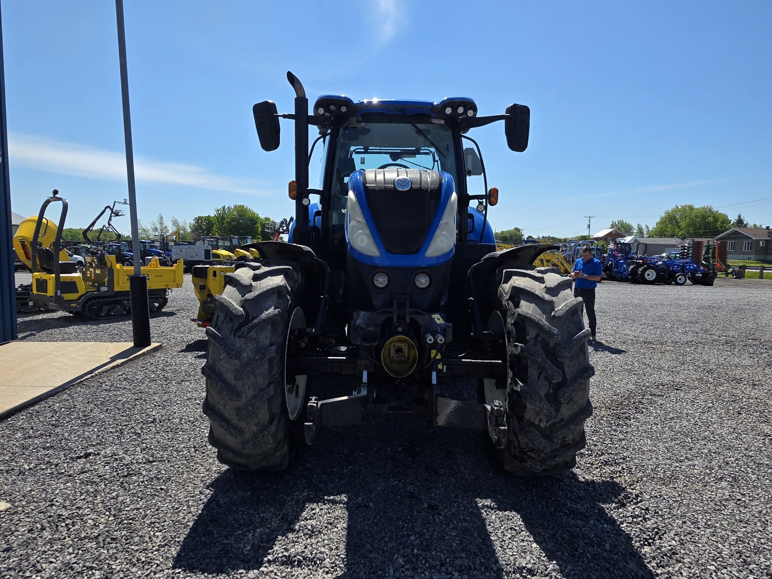 Un tracteur bleu est garé sur du gravier, face à la caméra, avec des engins de chantier et des personnes visibles en arrière-plan sous un ciel bleu clair.