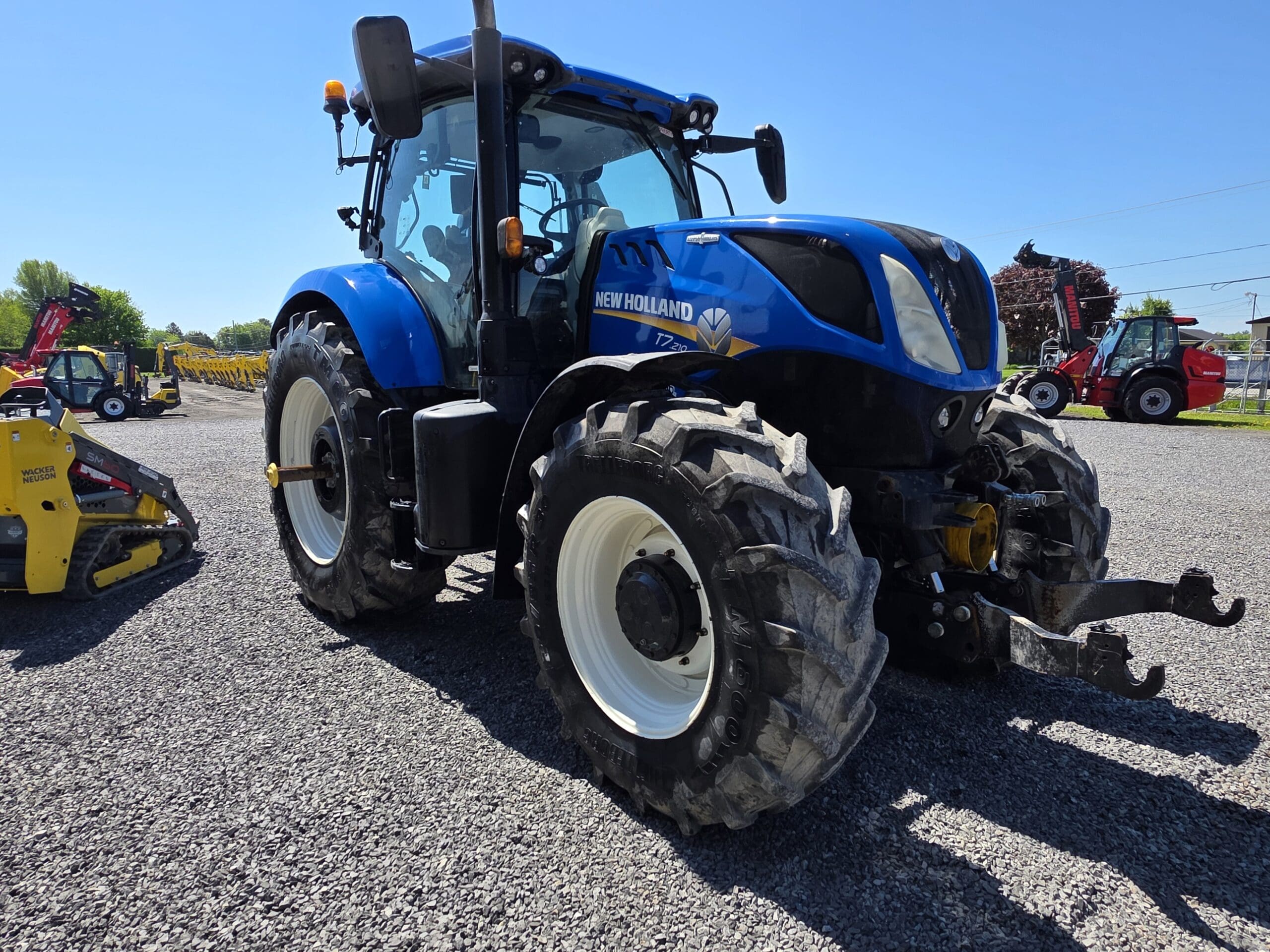 Un tracteur New Holland bleu est garé sur un terrain en gravier sous un ciel clair, avec d'autres équipements agricoles visibles en arrière-plan.
