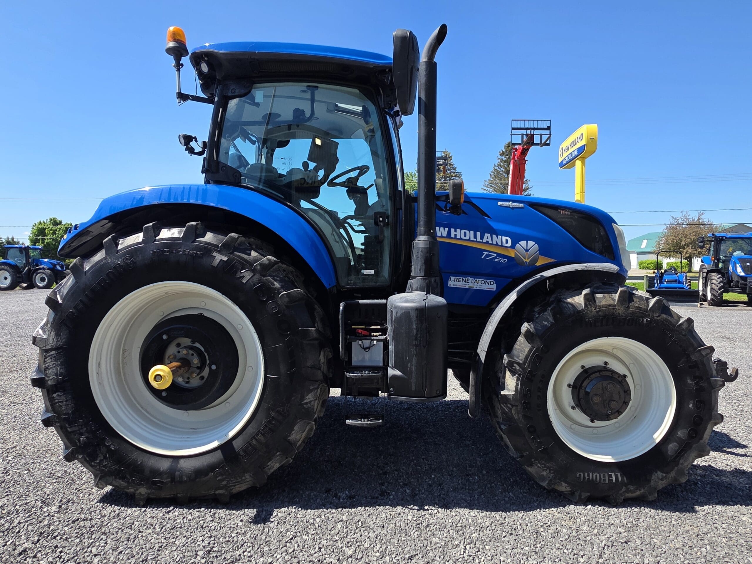 Un tracteur New Holland bleu garé sur du gravier, vu de côté avec des détails clairs de ses gros pneus arrière et de ses pneus avant plus petits sous un ciel bleu clair.