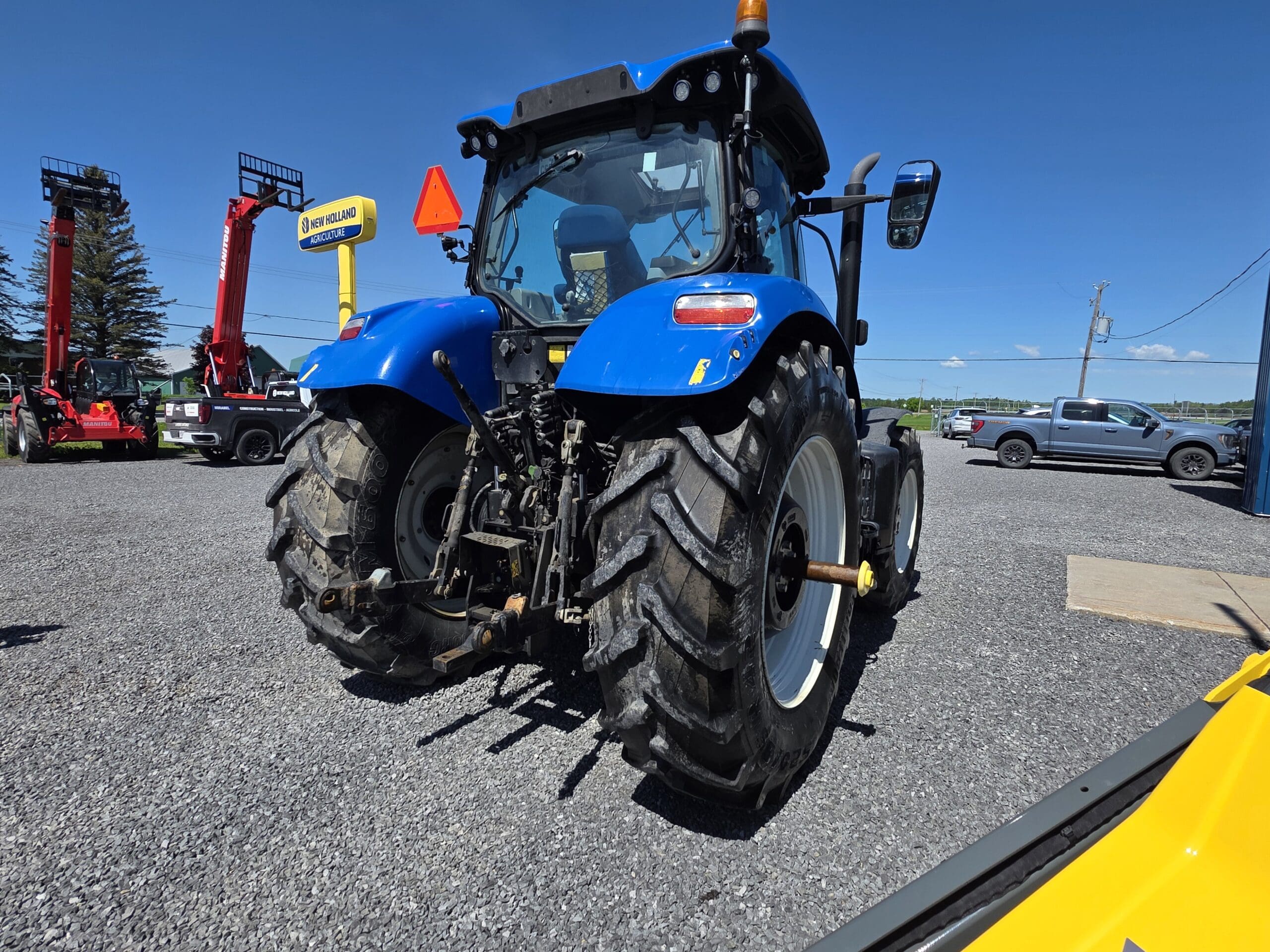 Un tracteur bleu est garé sur du gravier sous un ciel bleu clair, avec des véhicules et des équipements en arrière-plan, notamment un panneau de concessionnaire et un chariot élévateur rouge.