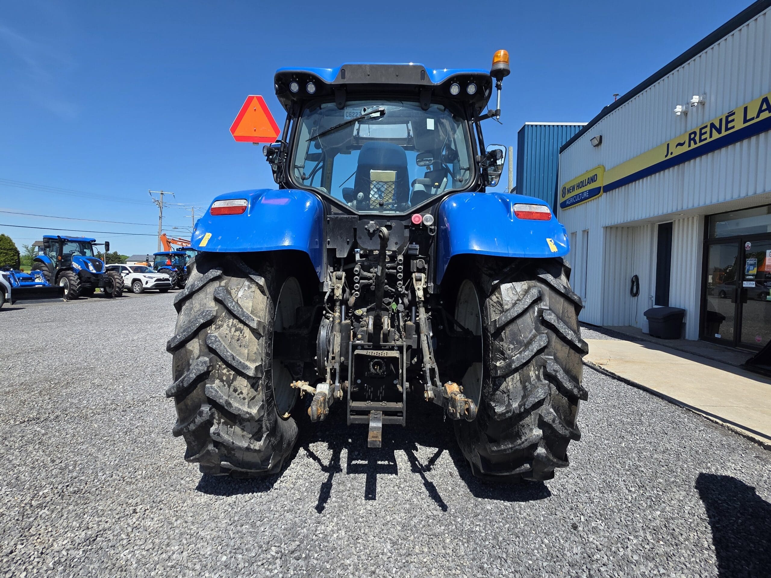 Un tracteur bleu est garé sur du gravier, vu de l'arrière. Il est équipé de gros pneus arrière robustes et de matériel agricole. Un bâtiment avec une enseigne se trouve à droite, et d'autres véhicules apparaissent en arrière-plan sous un ciel dégagé.
