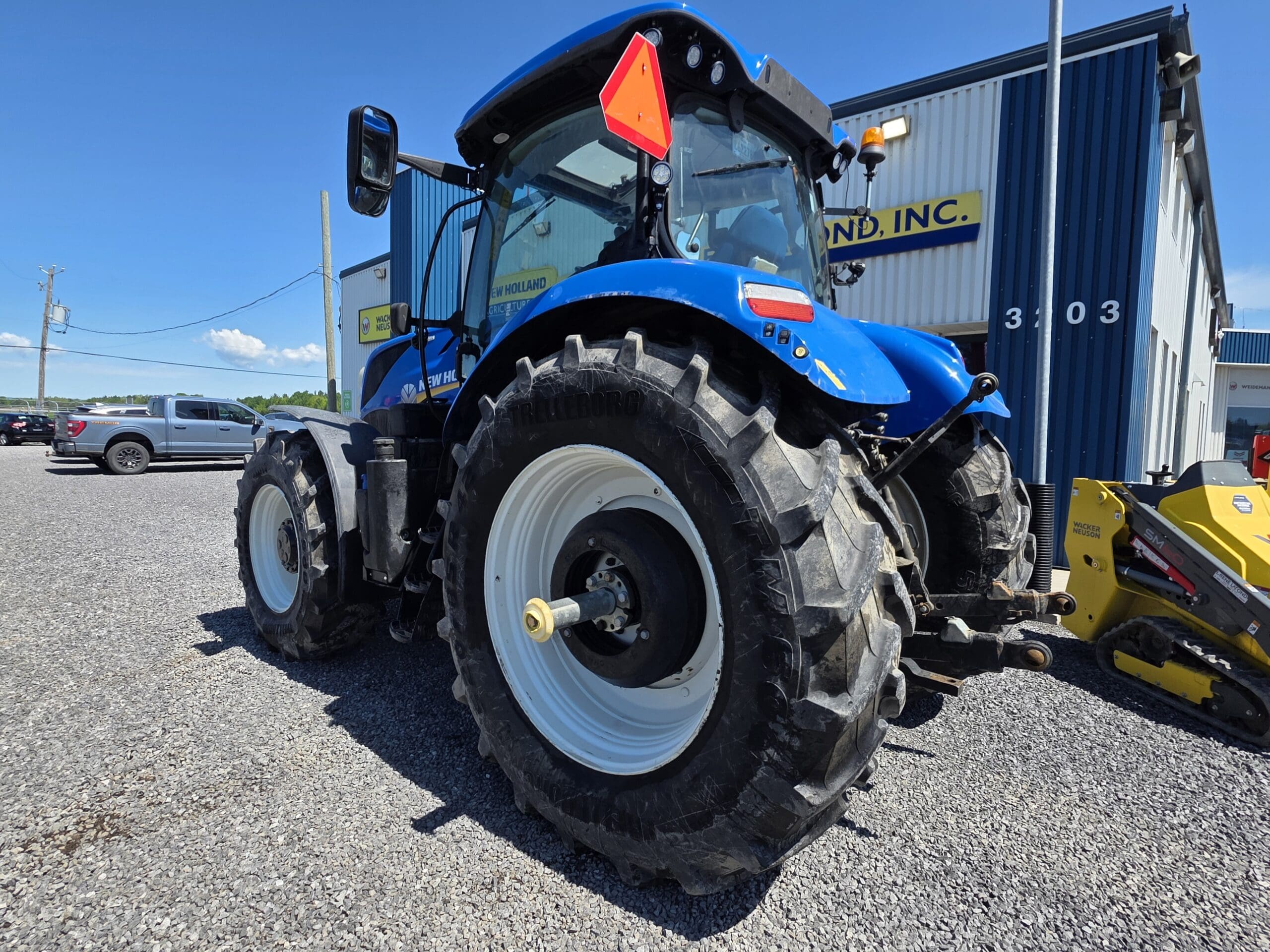 Un gros tracteur bleu aux pneus noirs est garé sur du gravier près d'un bâtiment métallique orné d'un panneau jaune. Le ciel est clair et ensoleillé.
