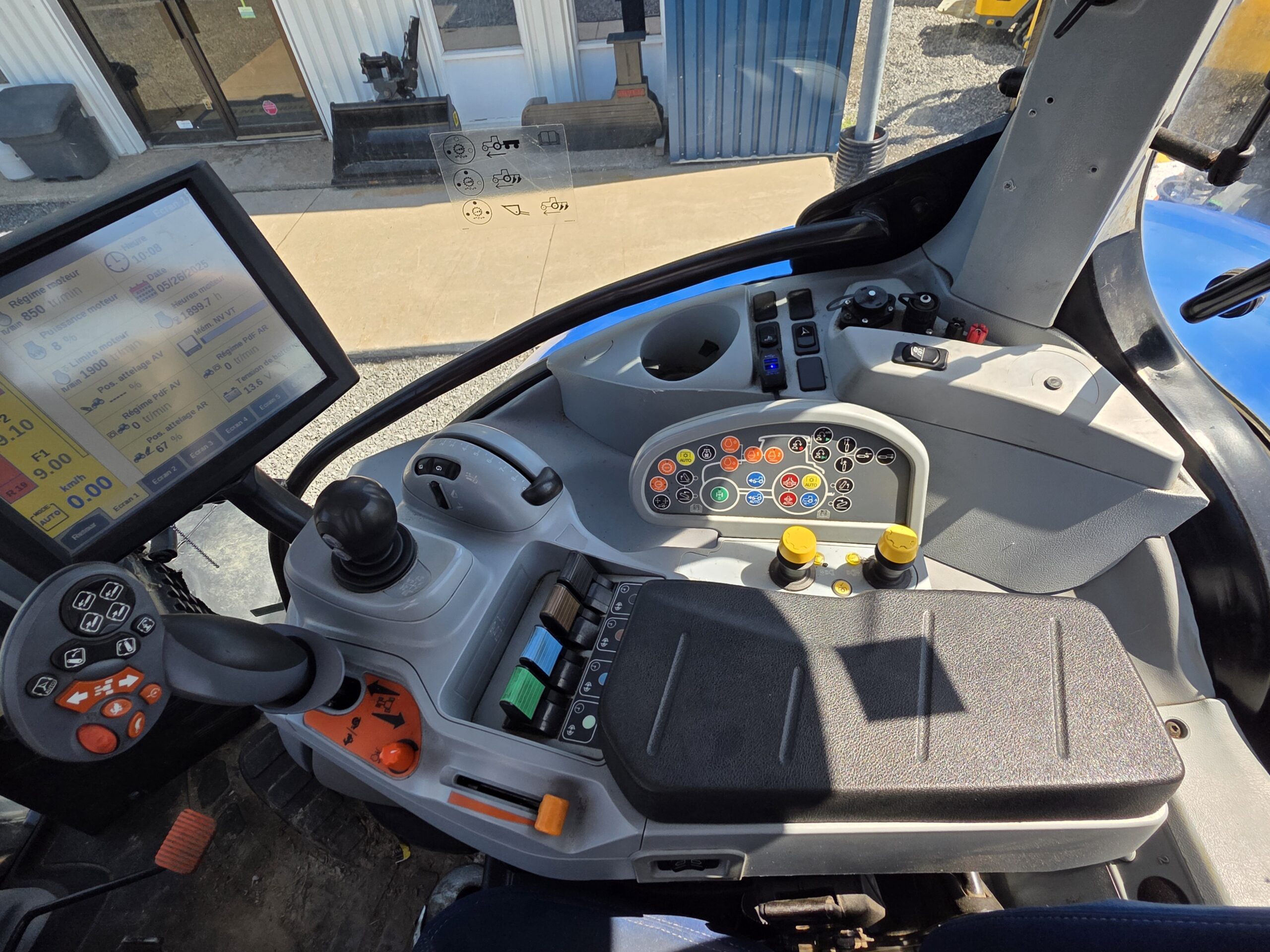 Panneau de commande et cockpit d'un tracteur moderne, montrant un écran tactile, un joystick, divers boutons, interrupteurs et leviers, avec une vue sur un extérieur industriel à travers la fenêtre de la cabine.