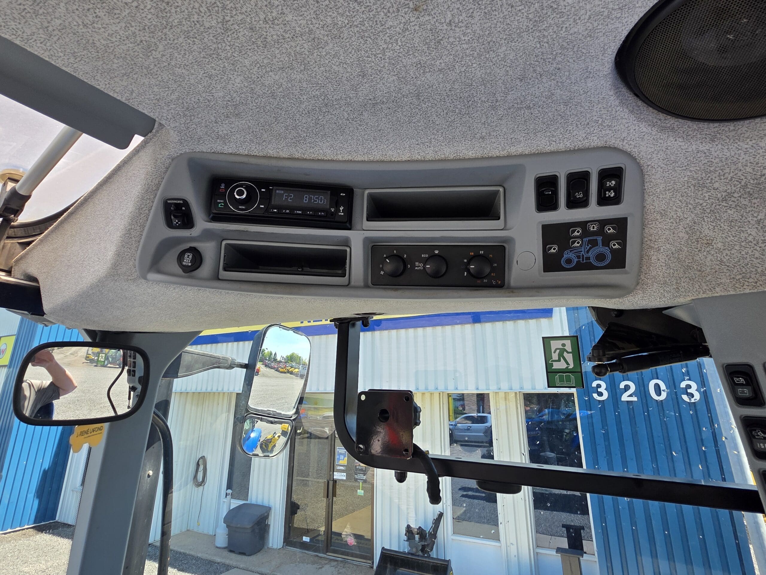 Vue du tableau de bord d'un tracteur avec radio, aérations, boutons et molettes. Les rétroviseurs latéraux et une partie du bâtiment bleu et blanc portant le numéro 3203 sont visibles à travers la fenêtre.