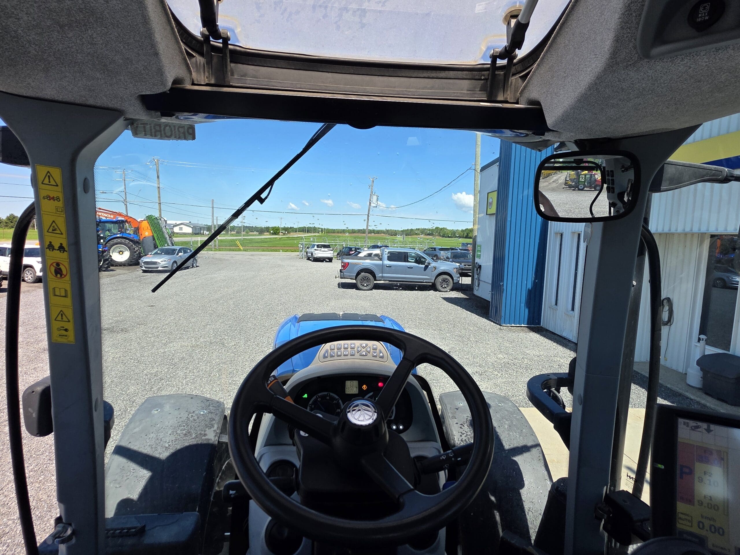 Vue depuis la cabine d'un tracteur, depuis le pare-brise, sur un parking en gravier avec plusieurs véhicules et un bâtiment. Les commandes du tracteur et le volant sont visibles au premier plan.