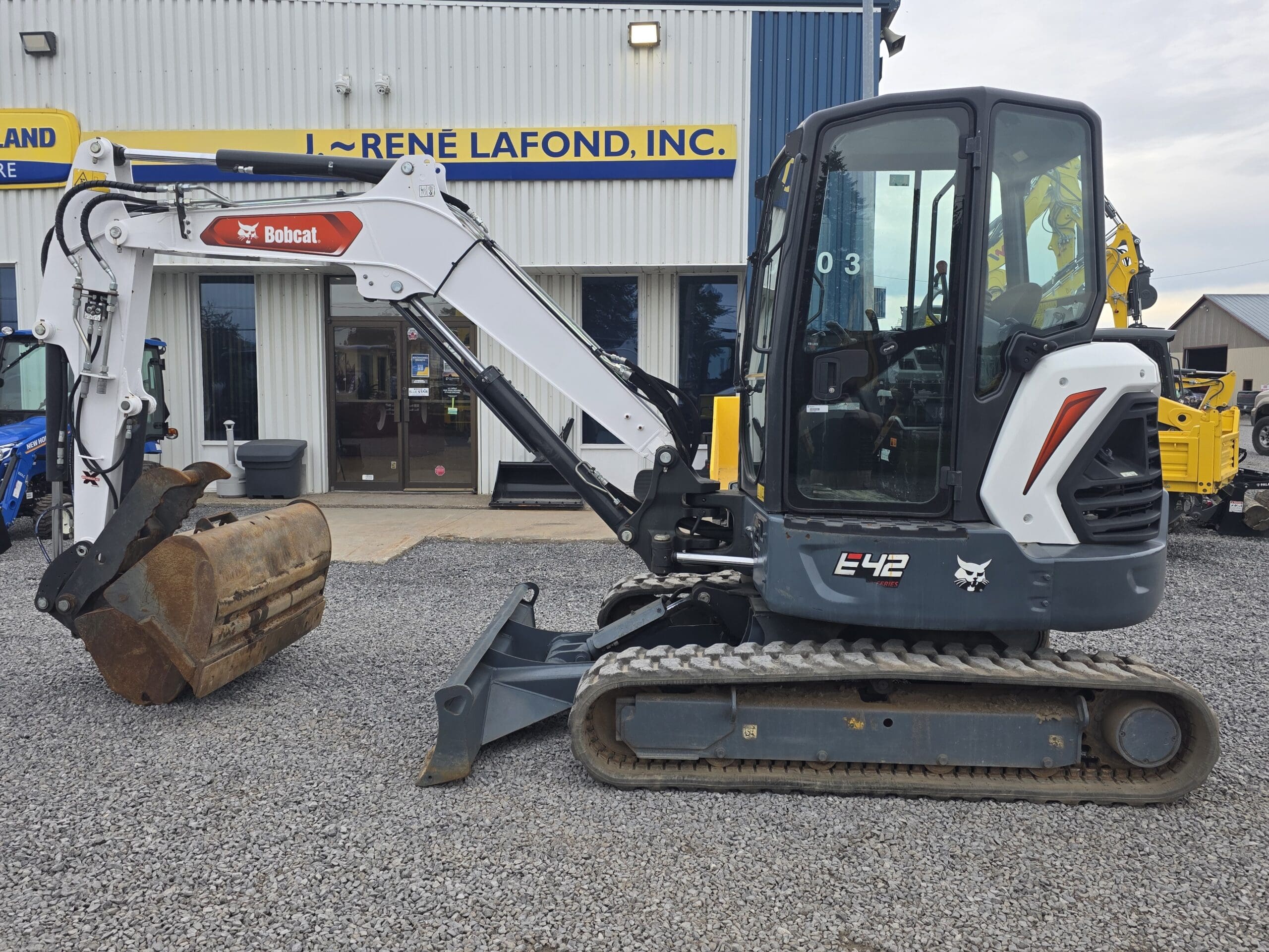 Une mini-pelle compacte Bobcat E42 blanche et noire avec un grand godet de creusement est garée sur du gravier à l'extérieur d'un bâtiment avec un panneau bleu et jaune indiquant « RENE LAFOND, INC.