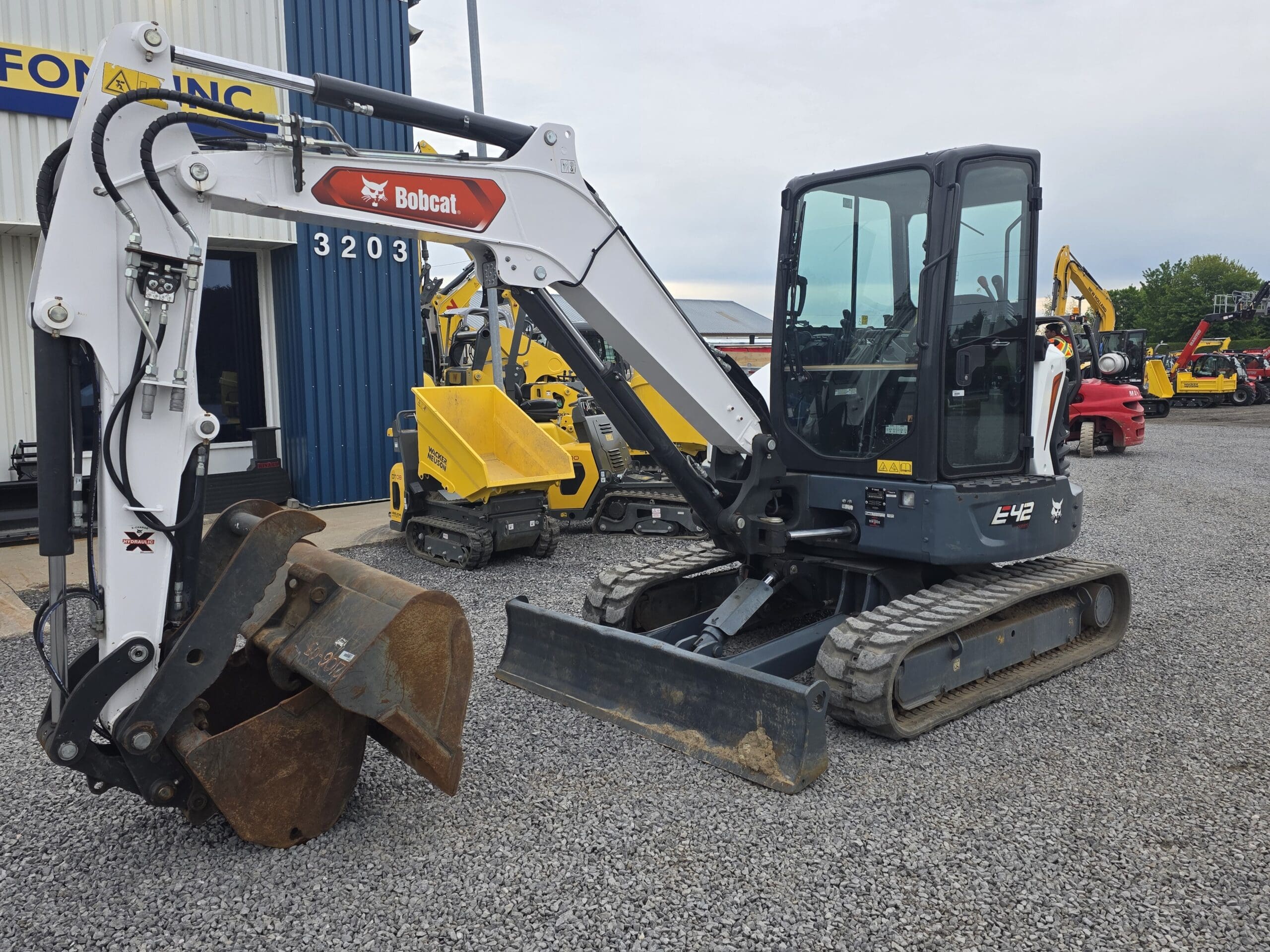 Une pelle compacte Bobcat E42 équipée d'un grand godet est garée sur du gravier à l'extérieur d'un bâtiment, avec d'autres engins de construction visibles en arrière-plan.