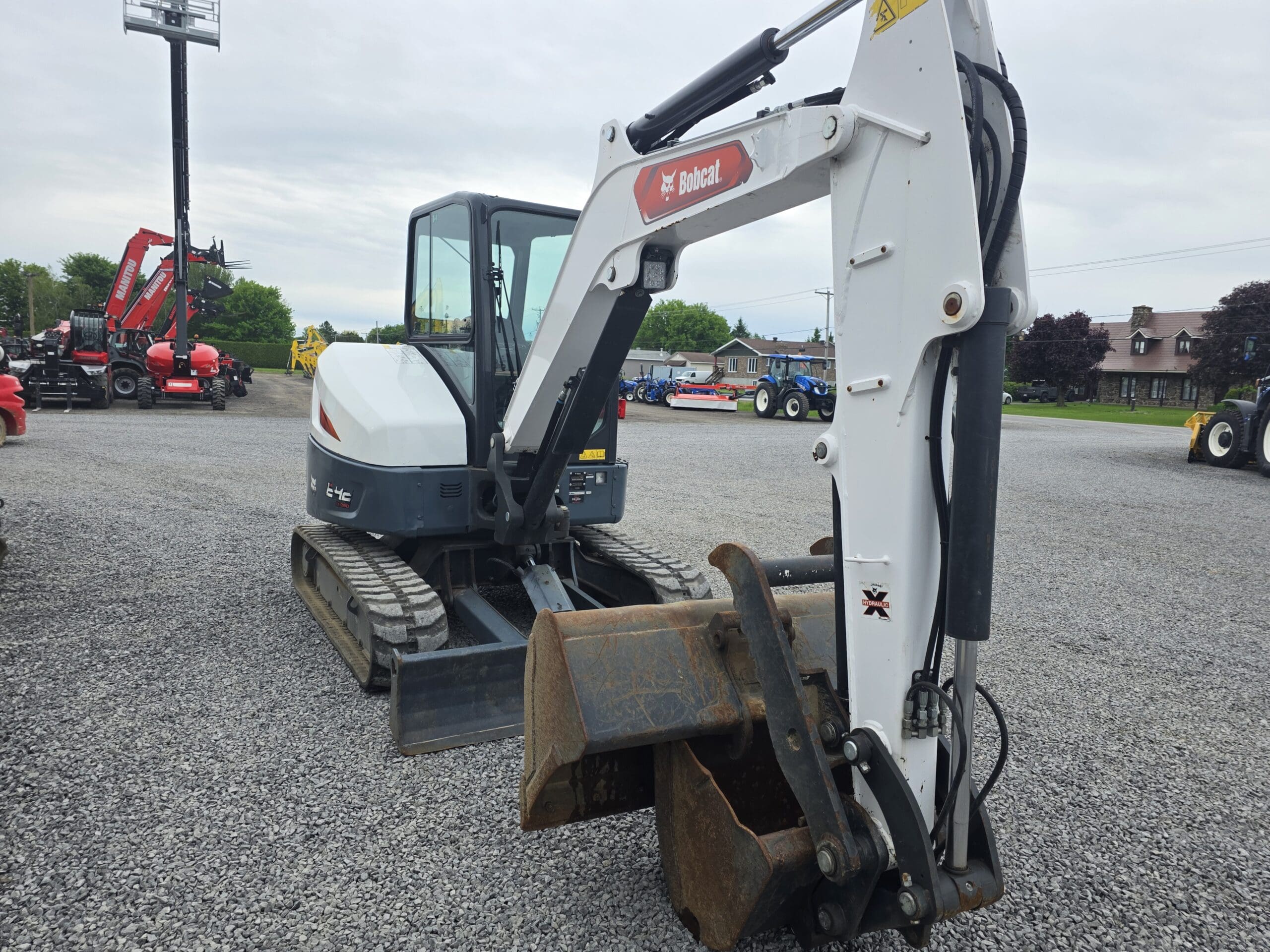 Une excavatrice compacte Bobcat blanche et noire avec un godet de creusement rouillé est garée sur un terrain en gravier, avec d'autres machines et arbres visibles en arrière-plan.