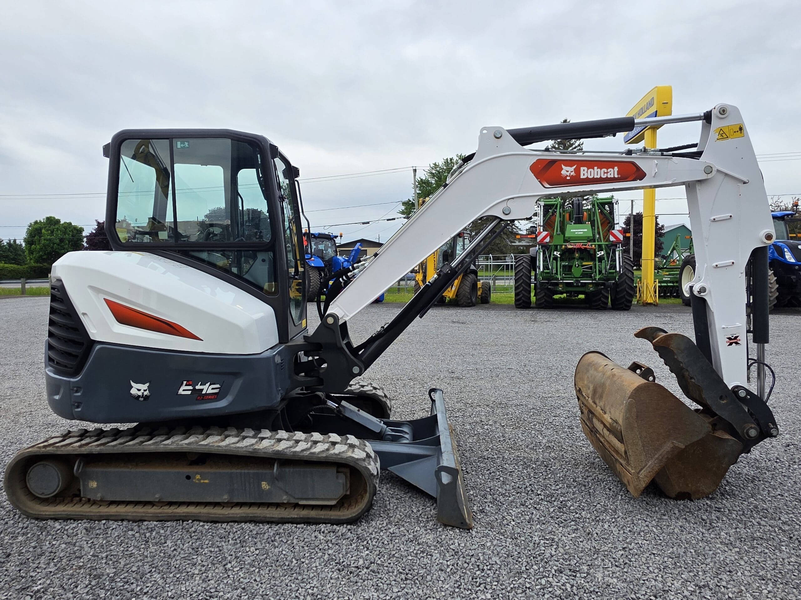 Excavatrice compacte Bobcat E42 blanche et grise avec une base à chenilles et un bras de creusement, garée sur du gravier à proximité d'autres machines agricoles sous un ciel nuageux.