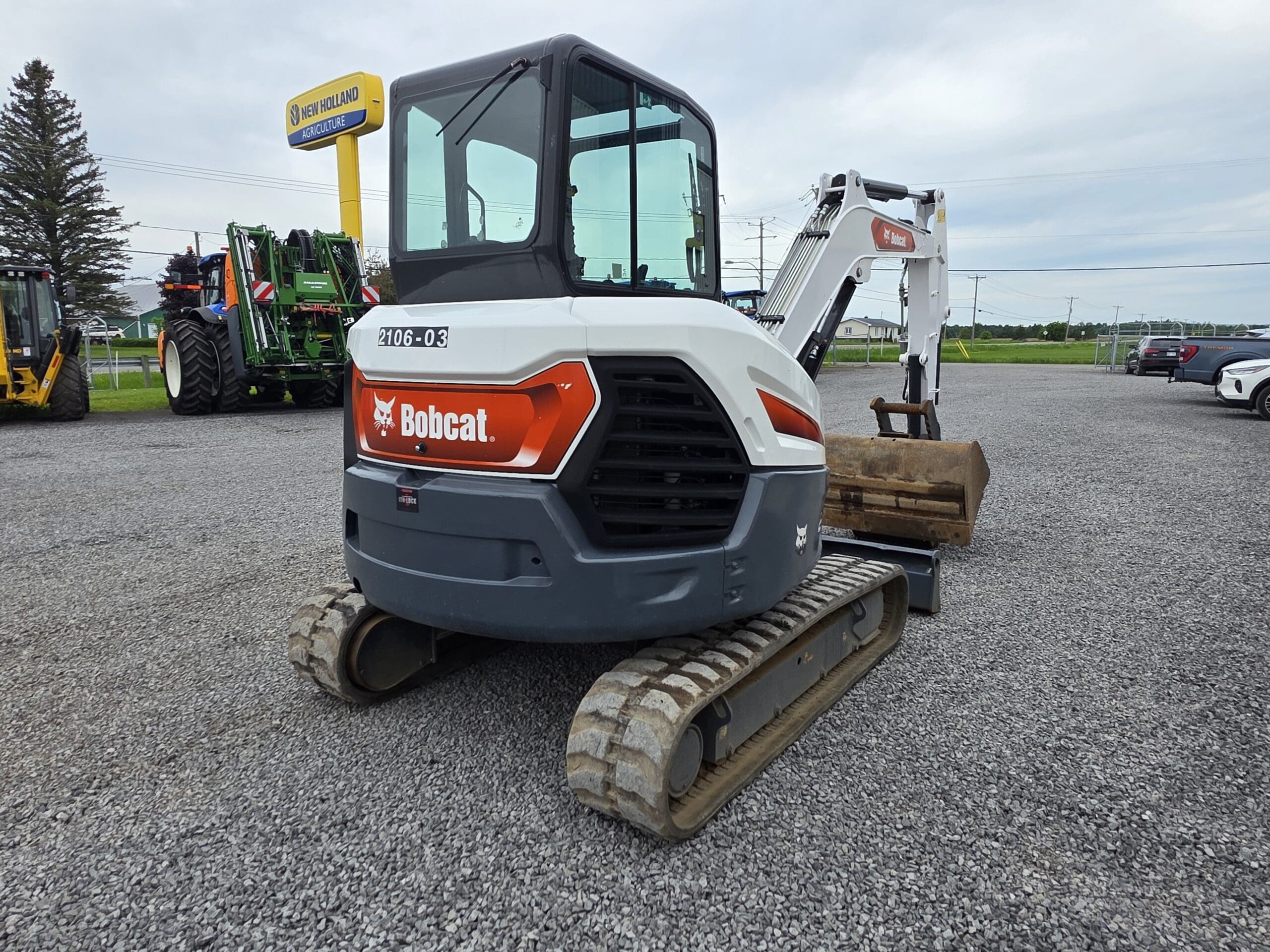 Une pelle compacte Bobcat blanche et orange est garée sur du gravier, équipée d'un godet en métal et de chenilles en caoutchouc. Des équipements agricoles et une enseigne New Holland sont visibles en arrière-plan.