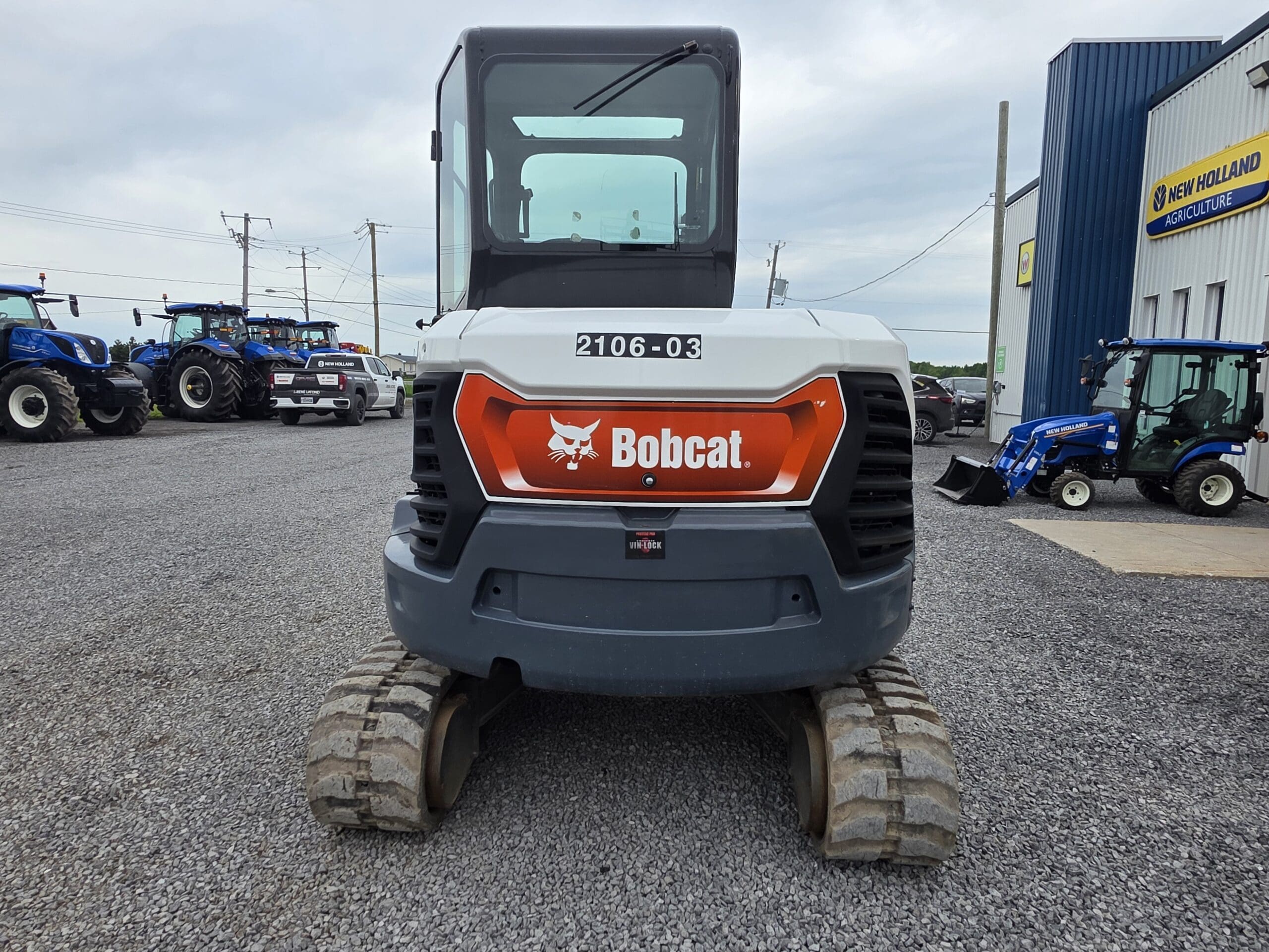 Une excavatrice Bobcat est garée sur du gravier, vue de l'arrière, avec la marque « Bobcat » visible. Des tracteurs et un bâtiment bleu portant l'inscription « New Holland Agriculture » sont visibles en arrière-plan.