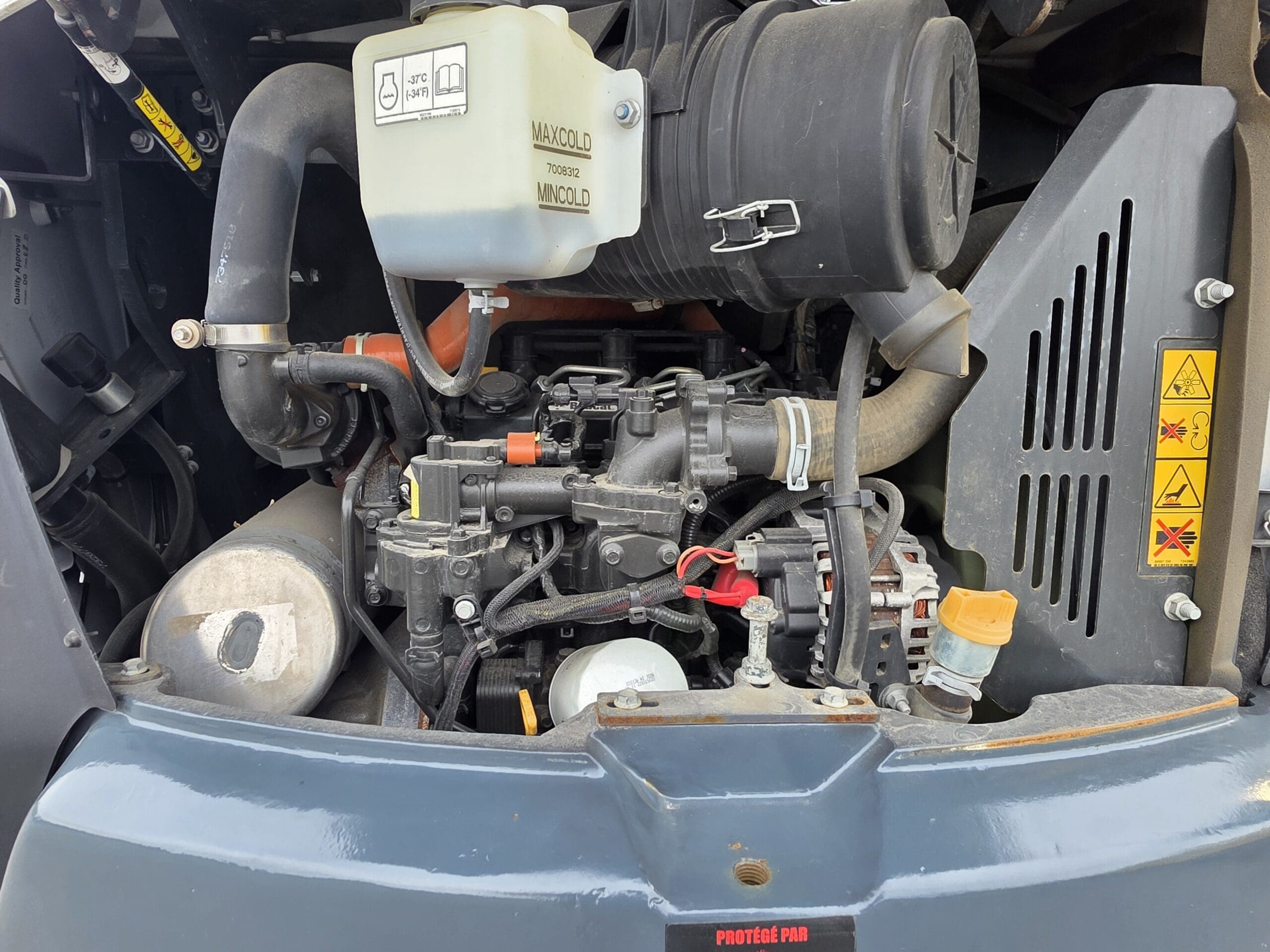 Gros plan du compartiment moteur d'un véhicule, montrant divers tuyaux, des fils, un réservoir de liquide de refroidissement et des autocollants d'avertissement sur une surface métallique. Les pièces du moteur semblent propres et bien rangées.