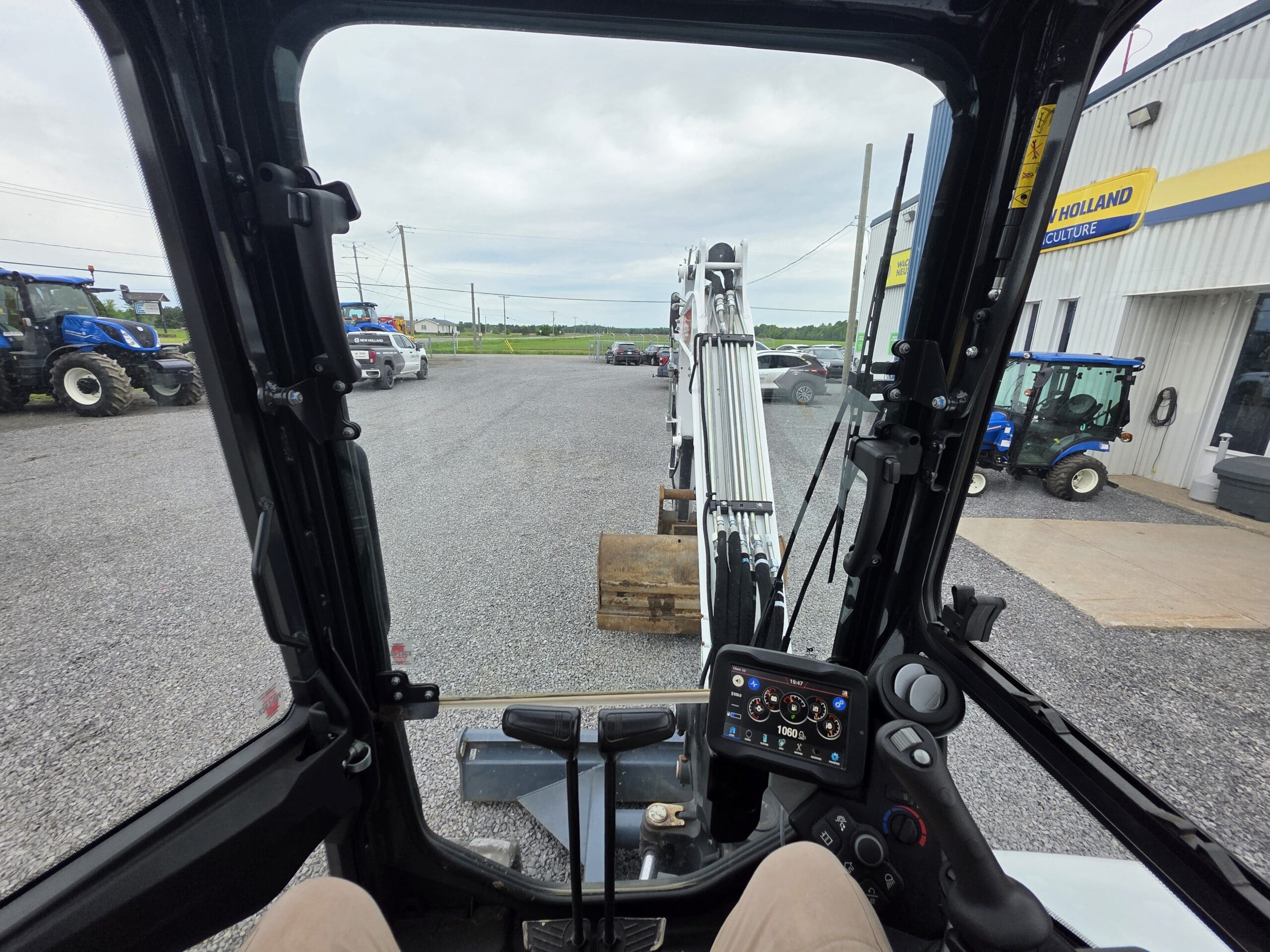 Vue depuis l'intérieur de la cabine d'un engin de chantier, avec vue sur un terrain gravillonné où se trouvent tracteurs et équipements. On peut voir les commandes du tableau de bord et le volant, ainsi que le bras avant et le godet de l'engin.