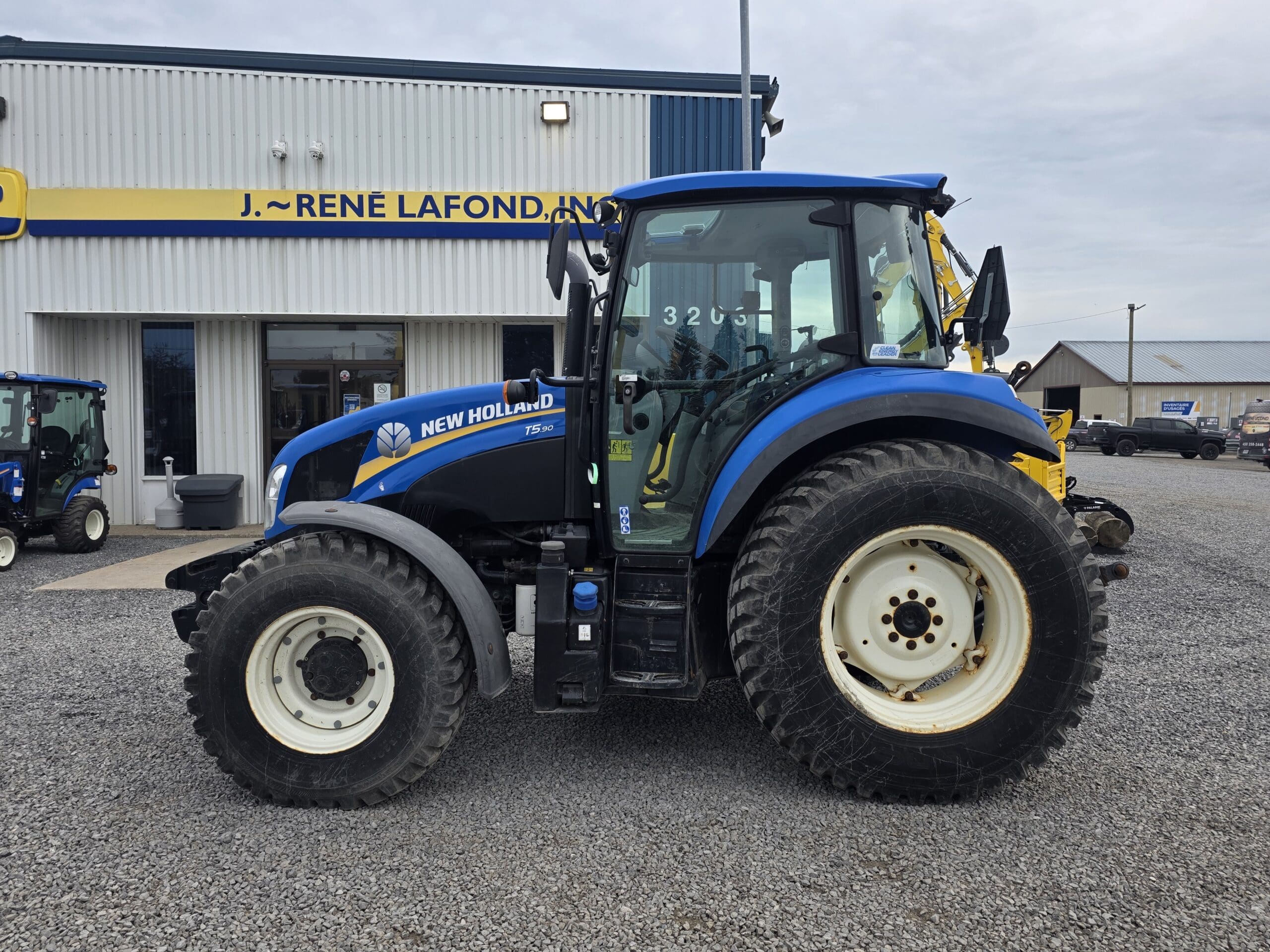 Un tracteur New Holland bleu est garé sur du gravier devant un bâtiment portant l'inscription « J. Rene Lafond, Inc. ». Le tracteur est vu de côté, avec de gros pneus et des vitres transparentes.
