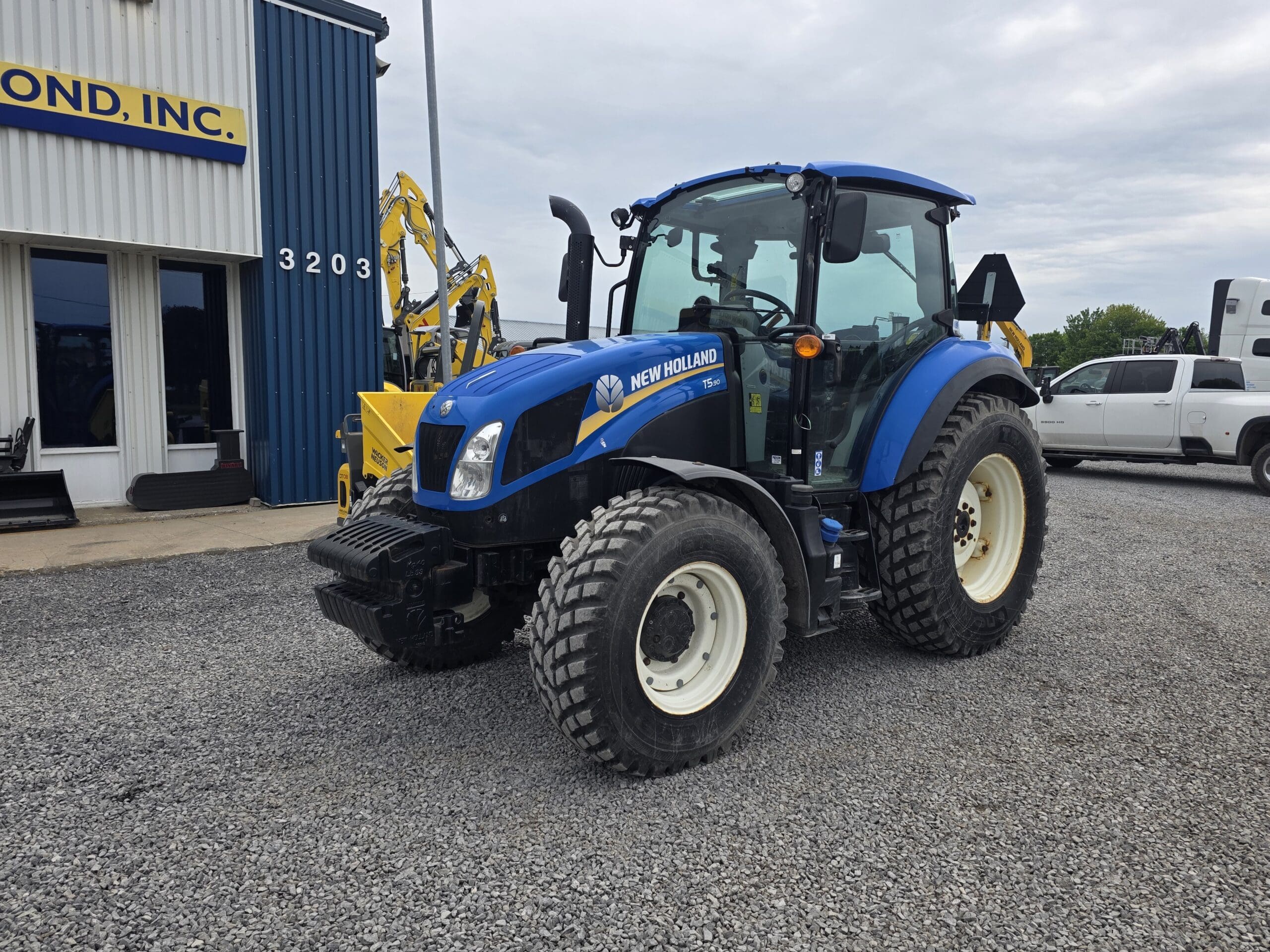 Un tracteur New Holland bleu est garé sur du gravier devant un bâtiment aux bardages bleus et blancs. Des équipements jaunes et des véhicules blancs sont visibles en arrière-plan.