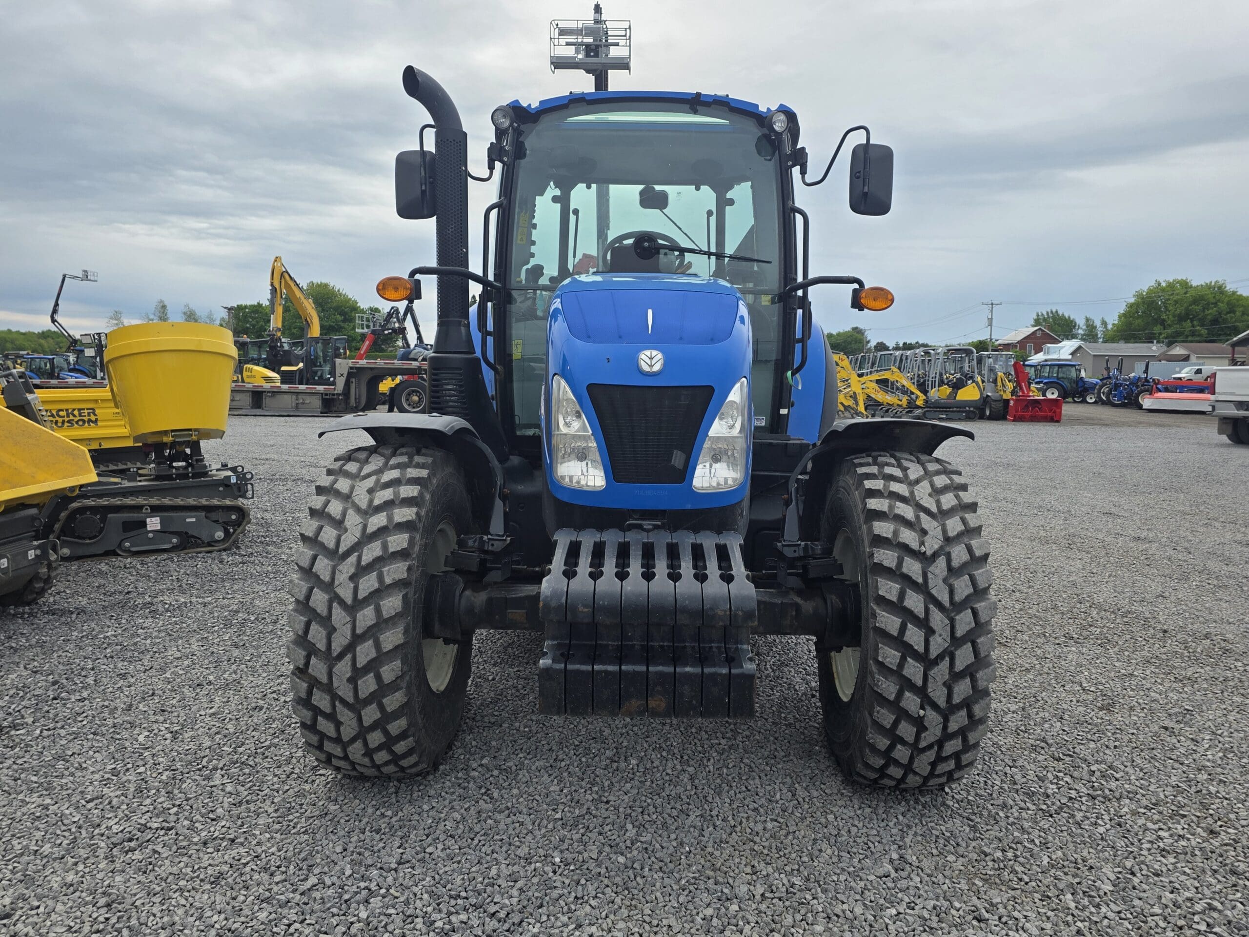 Un tracteur bleu garé sur du gravier avec des engins de chantier et des véhicules en arrière-plan sous un ciel nuageux.