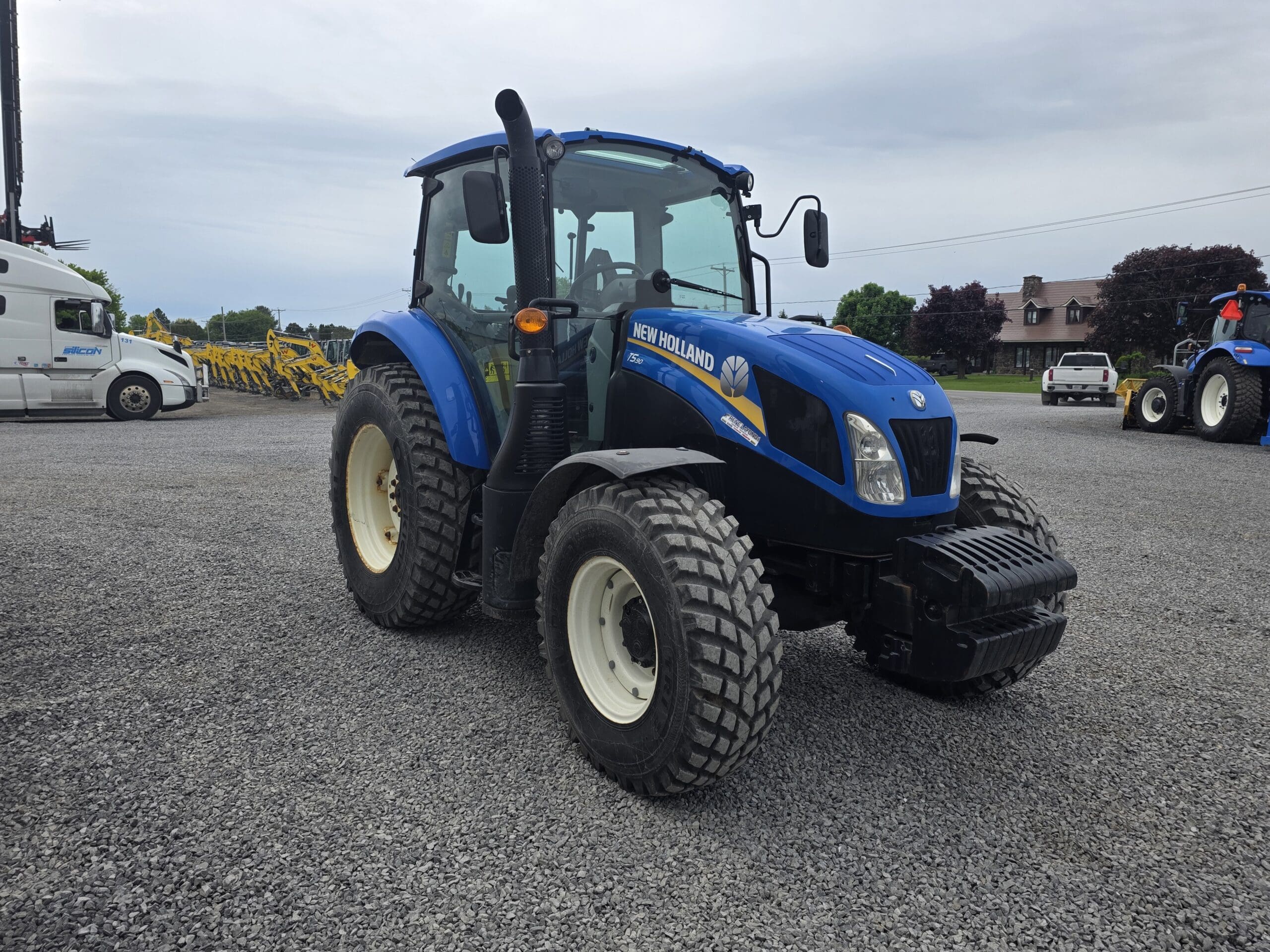 Un tracteur New Holland bleu est garé sur un terrain en gravier, avec un autre tracteur et une camionnette blanche visibles en arrière-plan sous un ciel nuageux.