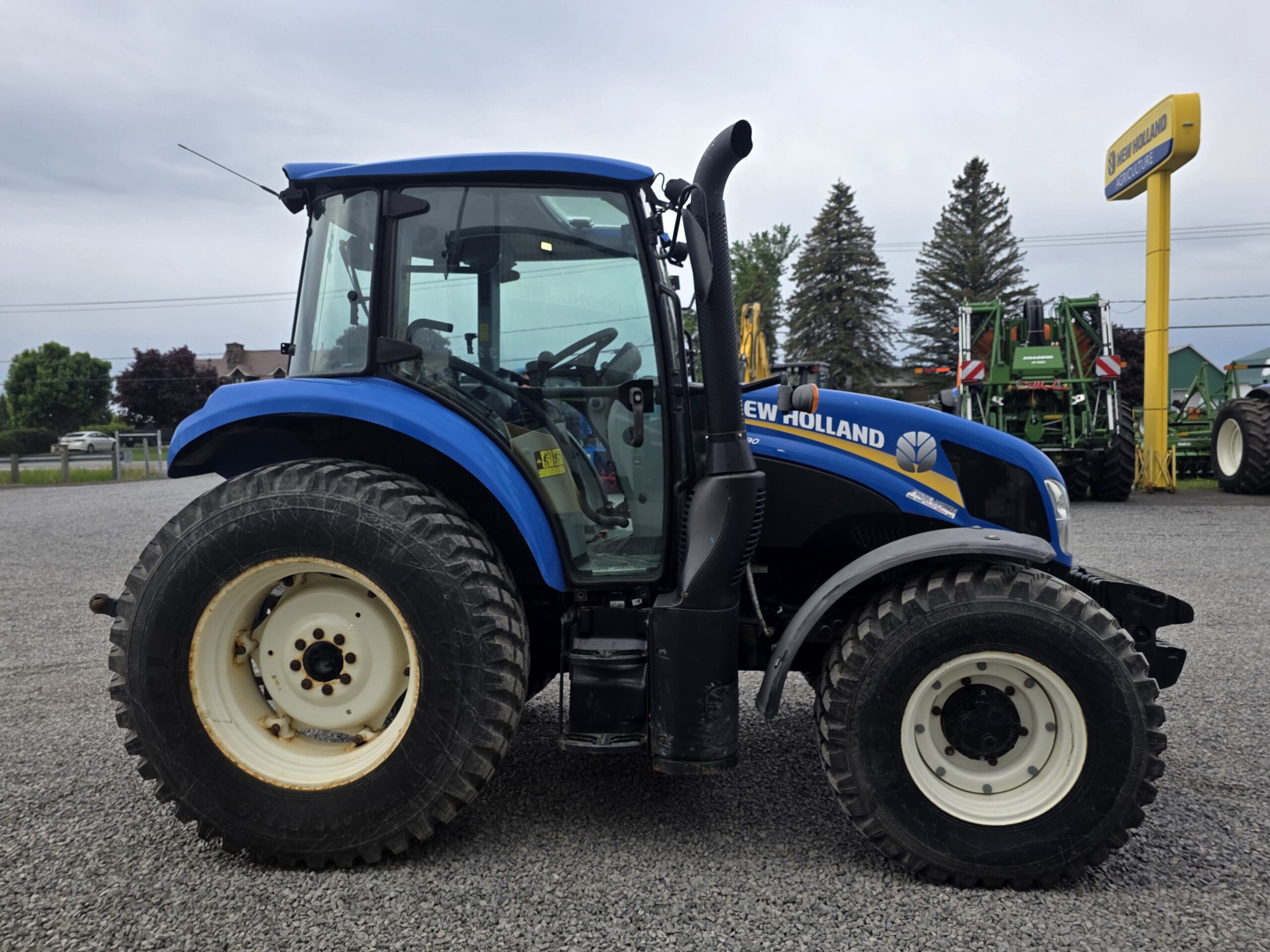Un tracteur New Holland bleu avec de gros pneus arrière et des pneus avant plus petits est garé sur un terrain en gravier à côté d'un panneau de concessionnaire jaune et de plusieurs pièces d'équipement agricole sous un ciel nuageux.