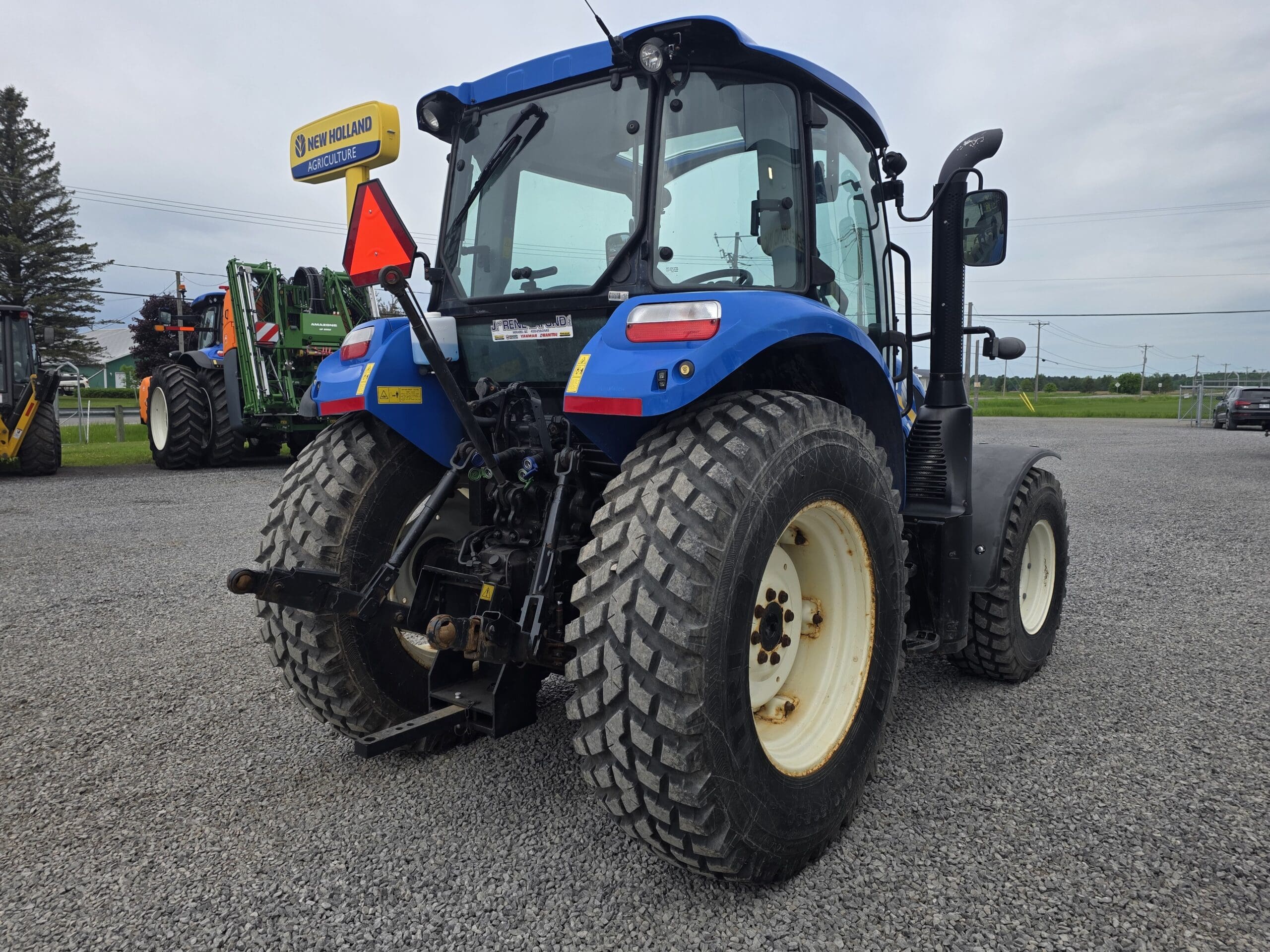 Un tracteur New Holland bleu est garé sur du gravier, vu de l'arrière. Il est équipé de gros pneus robustes et d'un panneau triangulaire de signalisation de véhicule lent à l'arrière. D'autres équipements et véhicules agricoles sont visibles en arrière-plan.