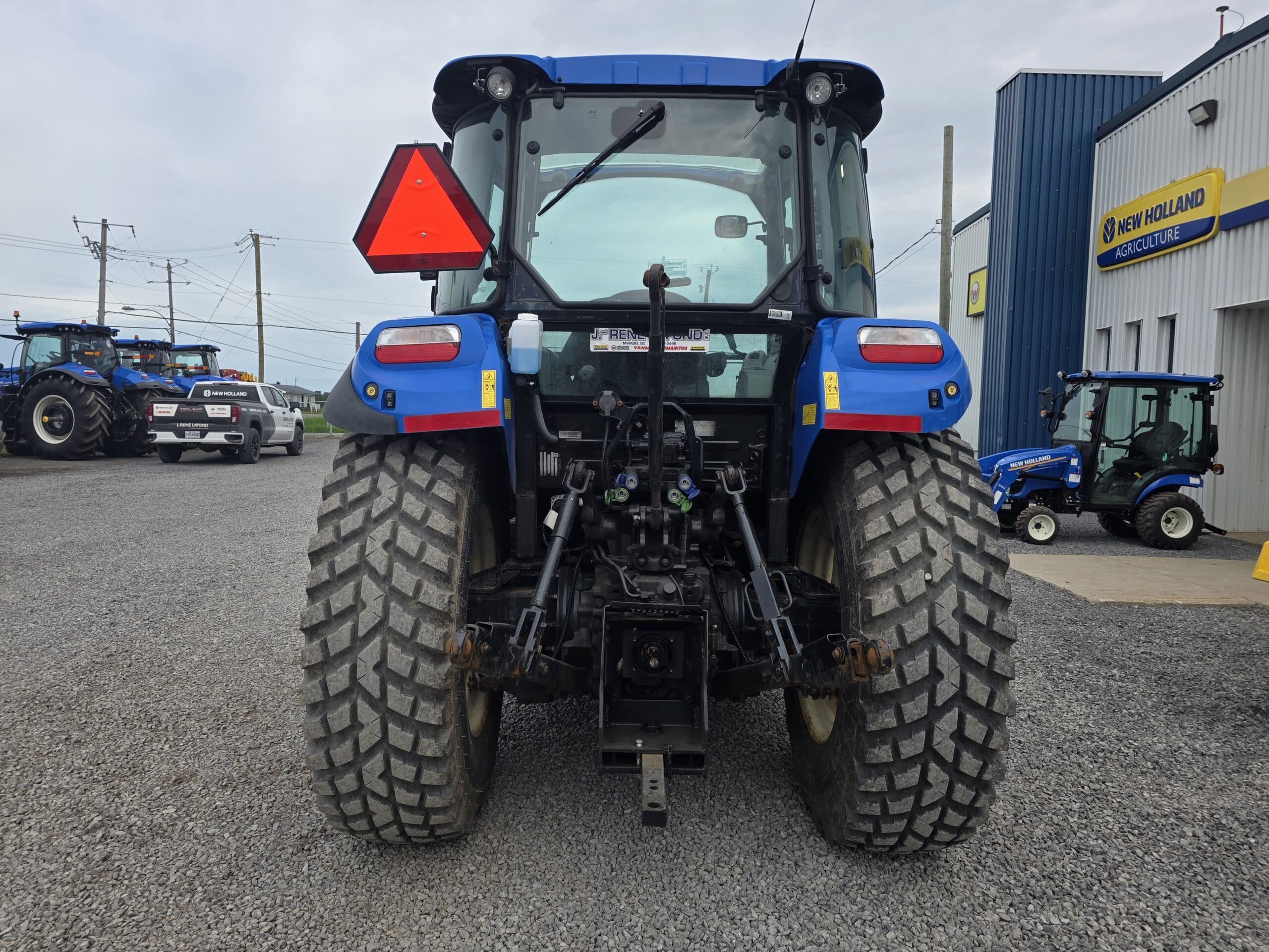 Vue arrière d'un tracteur bleu garé sur du gravier près d'un bâtiment agricole New Holland, avec un autre tracteur et des machines visibles en arrière-plan.