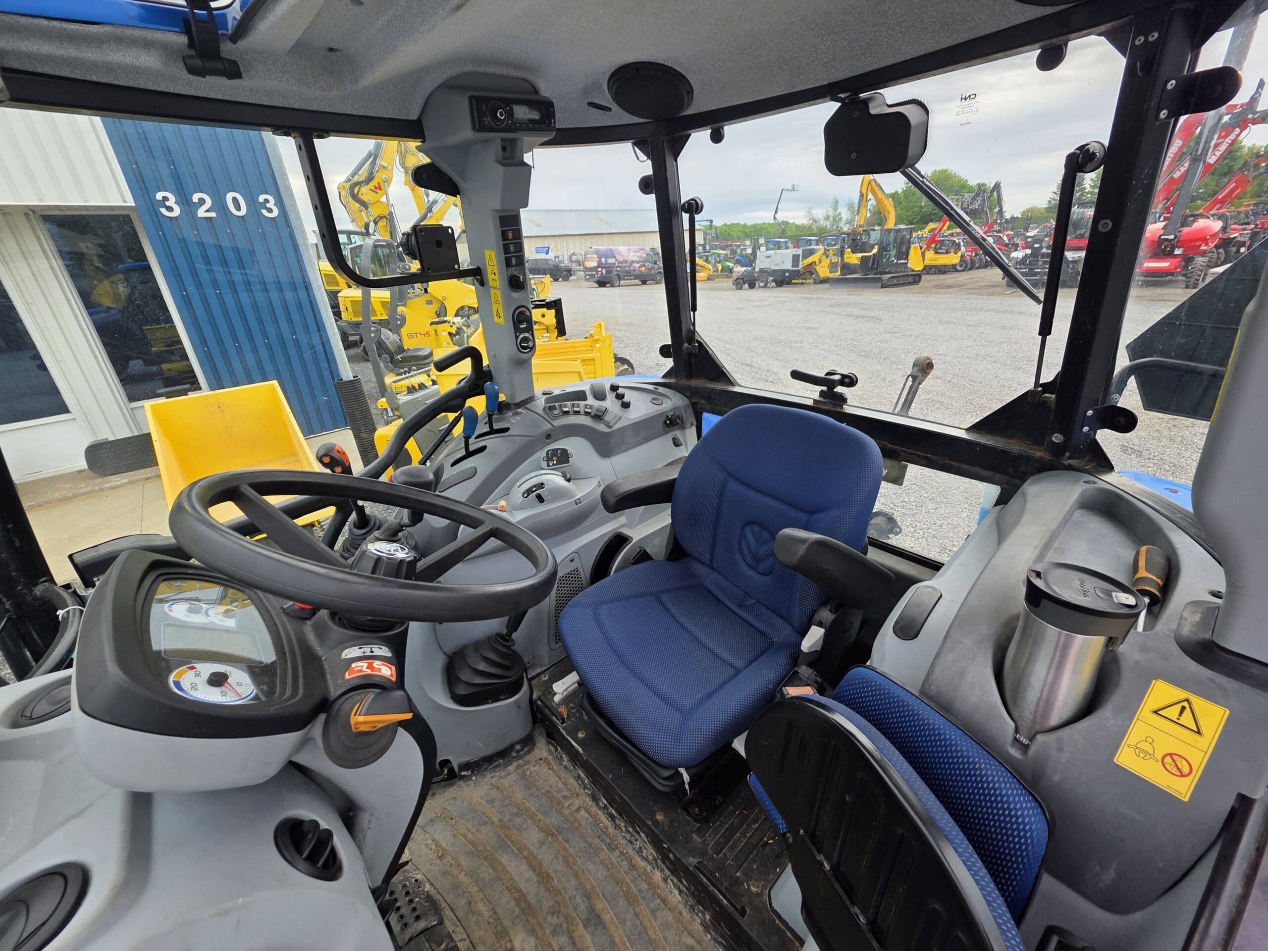 Vue intérieure d'une cabine de tracteur moderne, montrant le volant, les panneaux de commande, le siège conducteur bleu, divers leviers et le tableau de bord, avec l'équipement de construction visible à l'extérieur à travers les fenêtres.