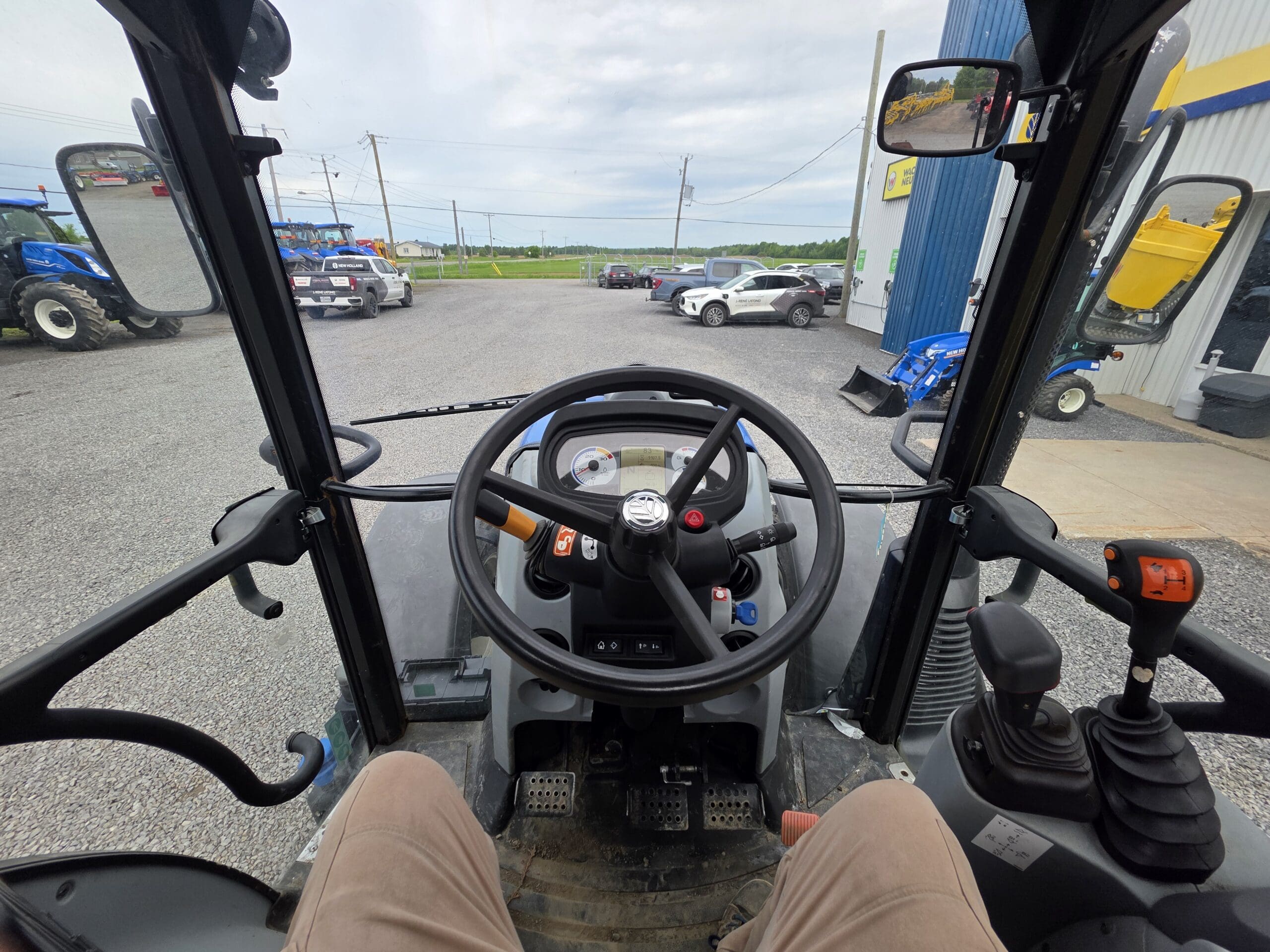 Vue de l'intérieur de la cabine d'un tracteur montrant le volant, les leviers de commande et les pédales. Le tracteur est garé à l'extérieur, avec d'autres véhicules et machines visibles sur un parking en gravier sous un ciel nuageux.