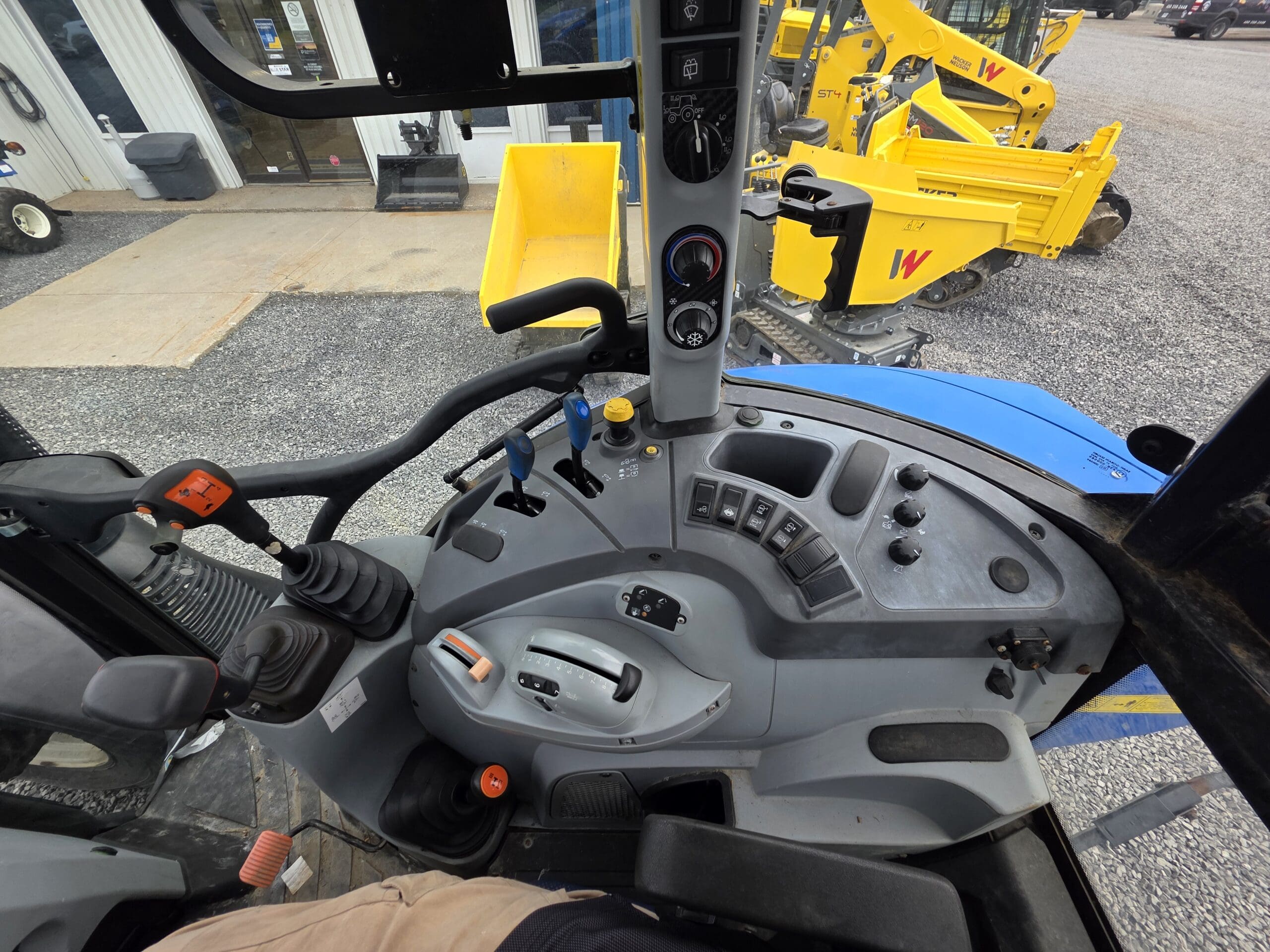 Vue de l'intérieur d'une cabine de tracteur montrant les leviers de commande, les boutons et les cadrans sur le tableau de bord, avec l'équipement de construction et le terrain de gravier visibles à travers la fenêtre extérieure.