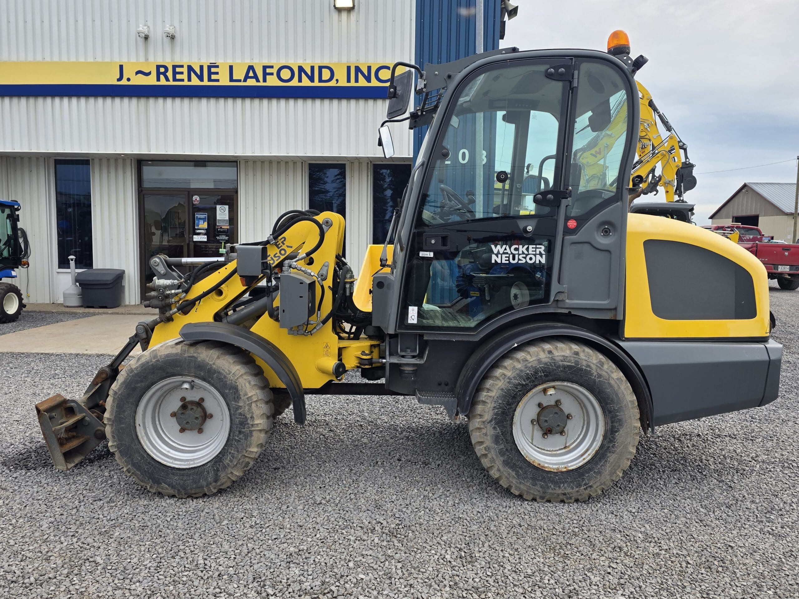 Une chargeuse compacte Wacker Neuson jaune et grise est garée sur du gravier devant un bâtiment portant une pancarte indiquant « J. Rene Lafond, Inc. » Une camionnette rouge est visible en arrière-plan.