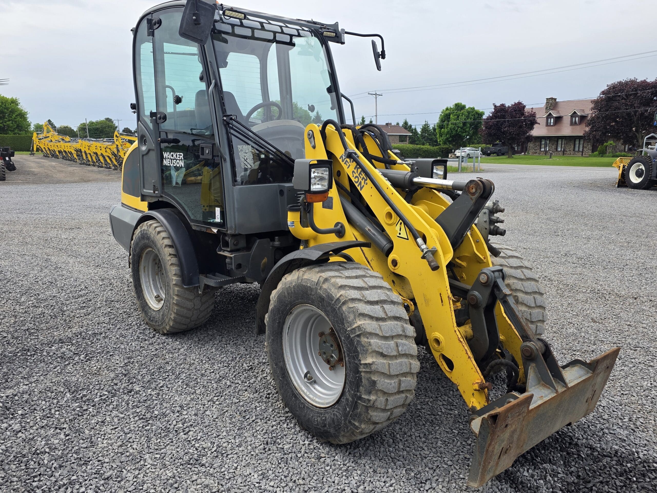 Une chargeuse compacte sur pneus jaune et noire avec un godet avant est garée sur un terrain en gravier, avec d'autres engins de construction et des arbres verts visibles en arrière-plan.