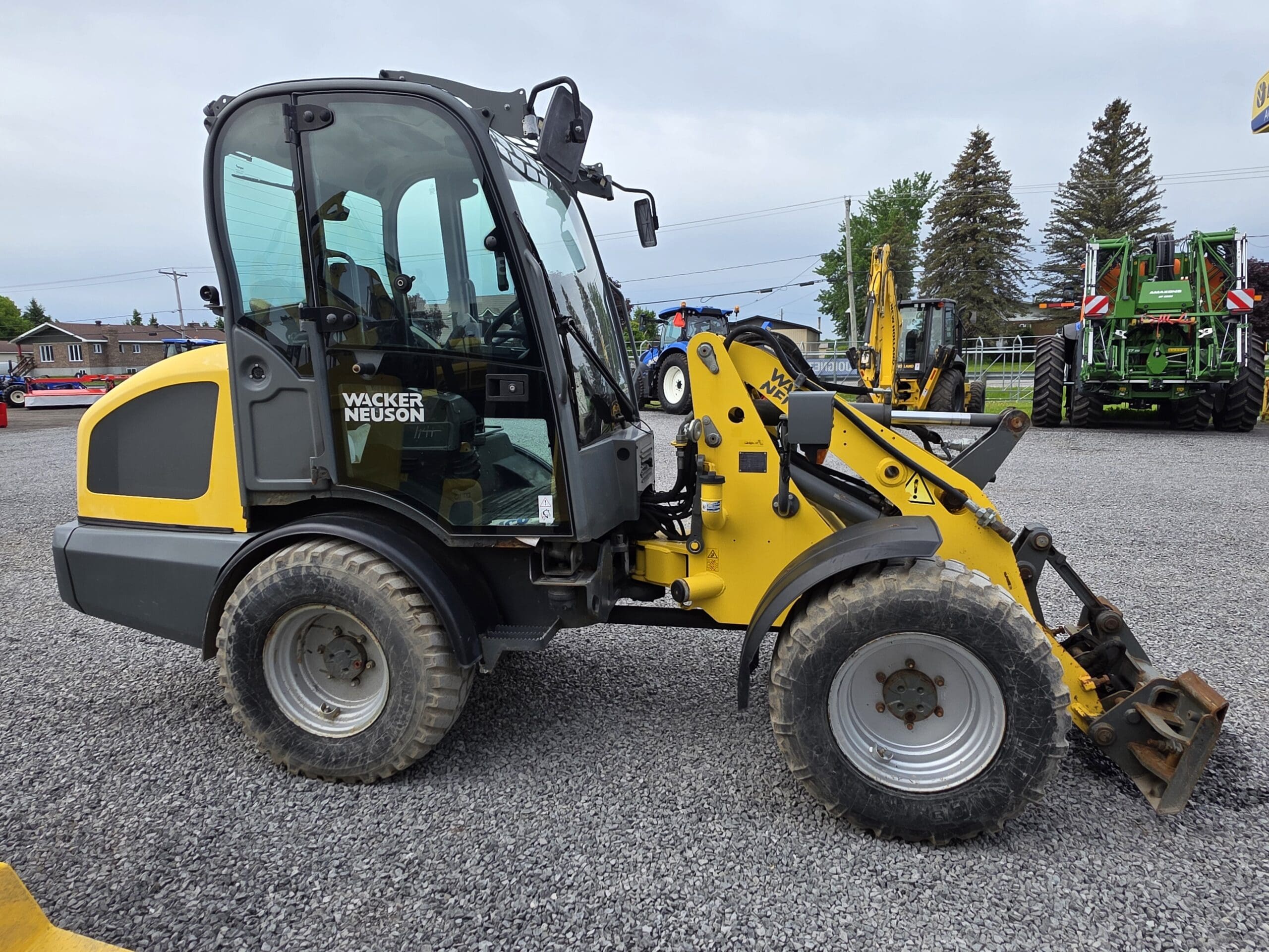 Une chargeuse compacte sur pneus Wacker Neuson jaune et noire est garée sur un terrain en gravier, avec d'autres machines et arbres visibles en arrière-plan sous un ciel nuageux.