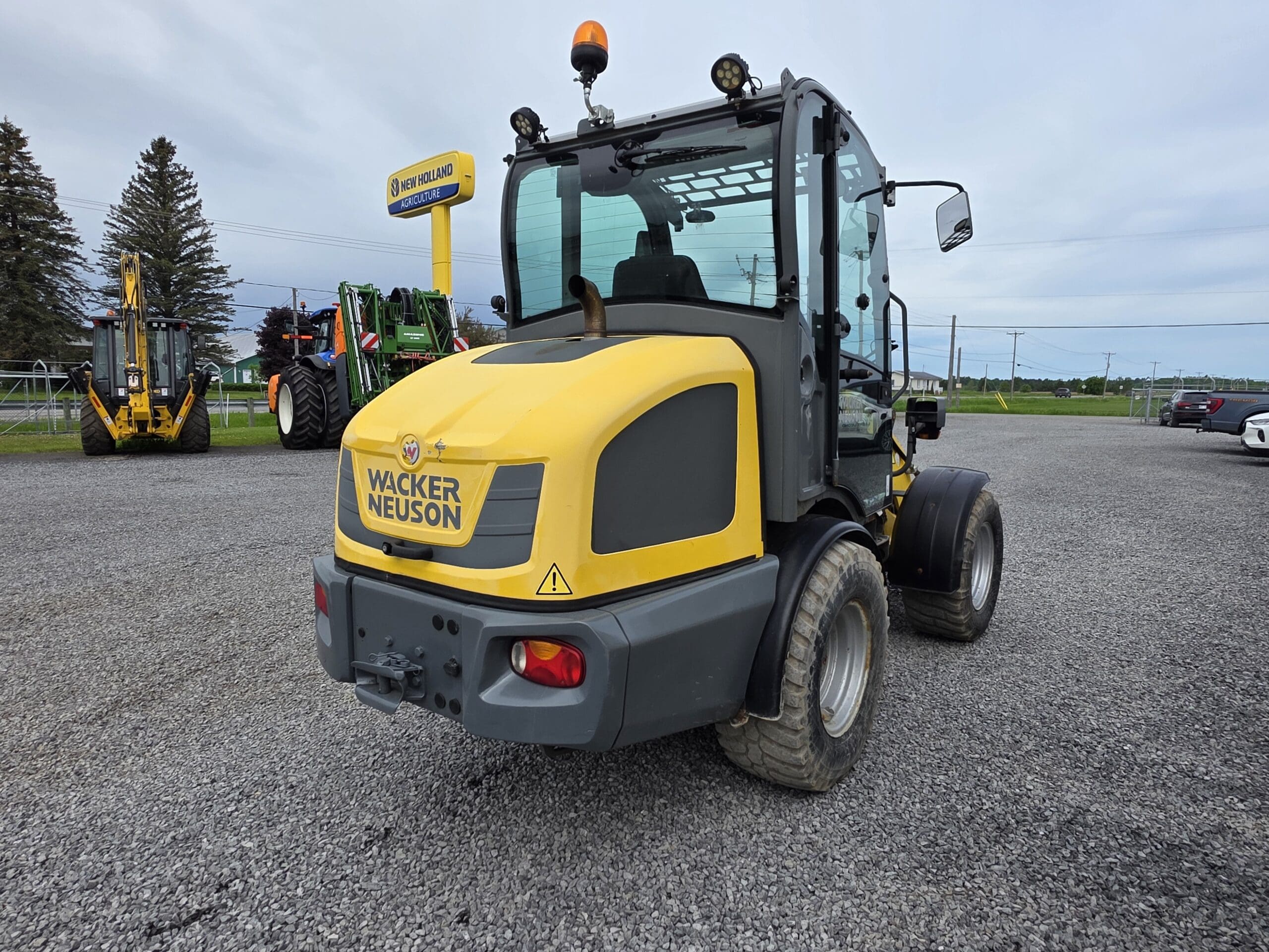 Un engin de chantier Wacker Neuson jaune est garé sur du gravier, vu de l'arrière. D'autres engins et véhicules sont visibles en arrière-plan sous un ciel nuageux.