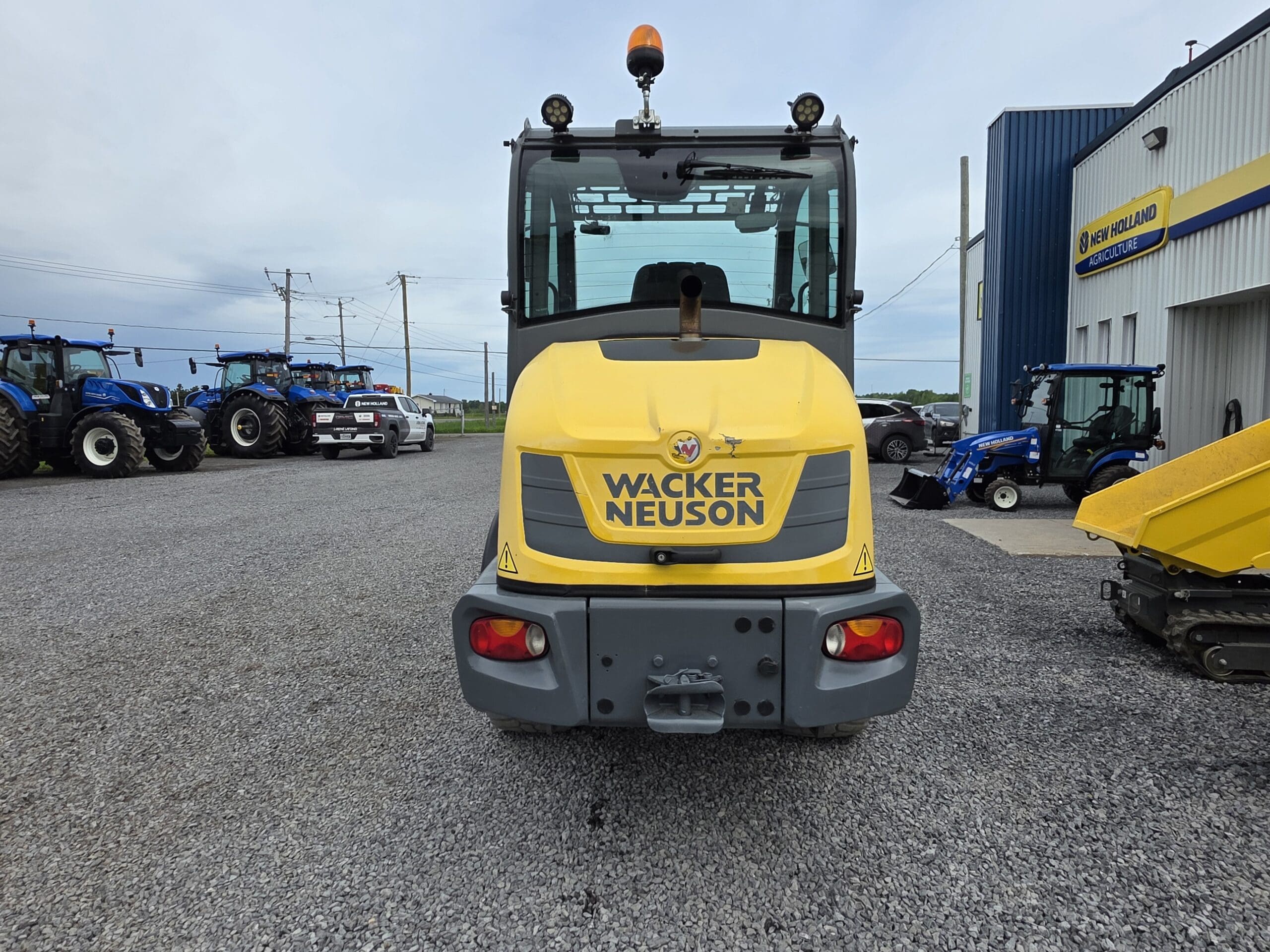 Un véhicule de construction jaune Wacker Neuson est garé sur du gravier à l'extérieur d'un concessionnaire, avec des tracteurs et des équipements visibles à gauche et à droite du véhicule.
