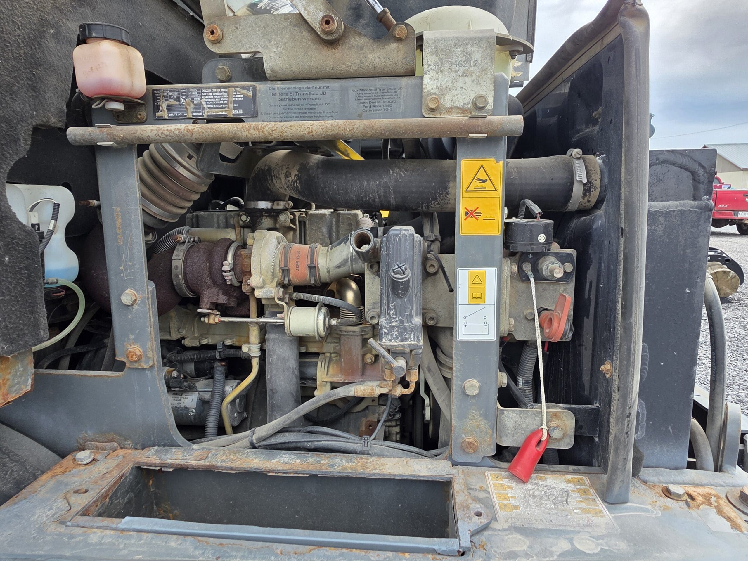 Gros plan sur les composants d'un moteur, notamment les tuyaux, les câbles, les étiquettes d'avertissement et un réservoir de liquide de refroidissement, avec des pièces métalliques apparentes et de la rouille visible. L'arrière-plan montre un ciel partiellement nuageux et un sol en gravier.