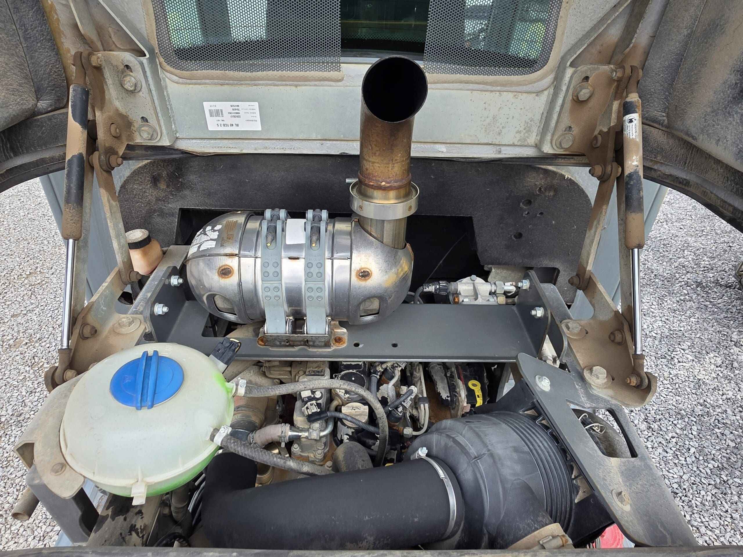 Gros plan sur le compartiment moteur d'une machine avec un gros tuyau d'échappement, divers flexibles, un réservoir de liquide de refroidissement avec un bouchon bleu et une structure métallique. Le moteur est visible sous un capot de protection.