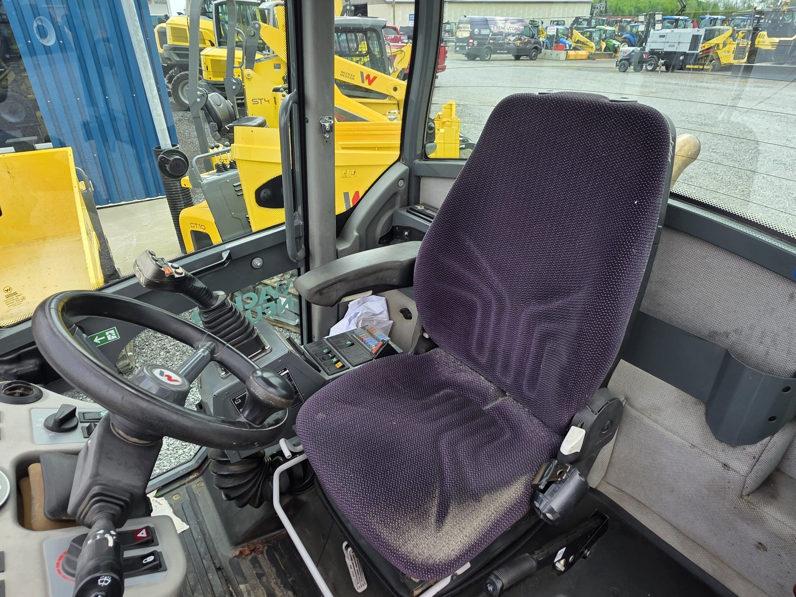 Siège conducteur et zone de commande à l'intérieur d'une cabine de véhicule de construction, comprenant un siège en tissu avec accoudoirs, un volant et un panneau de commande, avec des équipements de construction supplémentaires visibles à l'extérieur à travers les fenêtres.