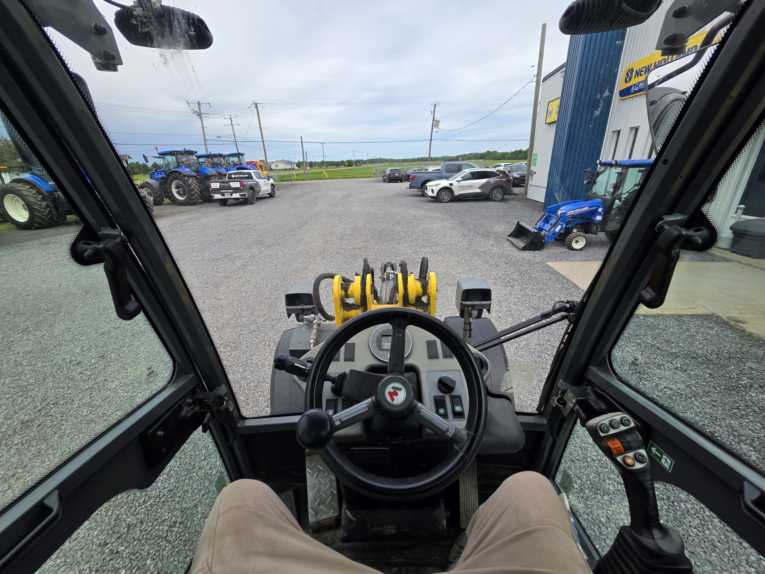 Vue de l'intérieur de la cabine d'un engin de chantier montrant le volant, le panneau de commande et les équipements extérieurs. À l'extérieur, on distingue des tracteurs, une voiture et un bâtiment bleu par temps nuageux.