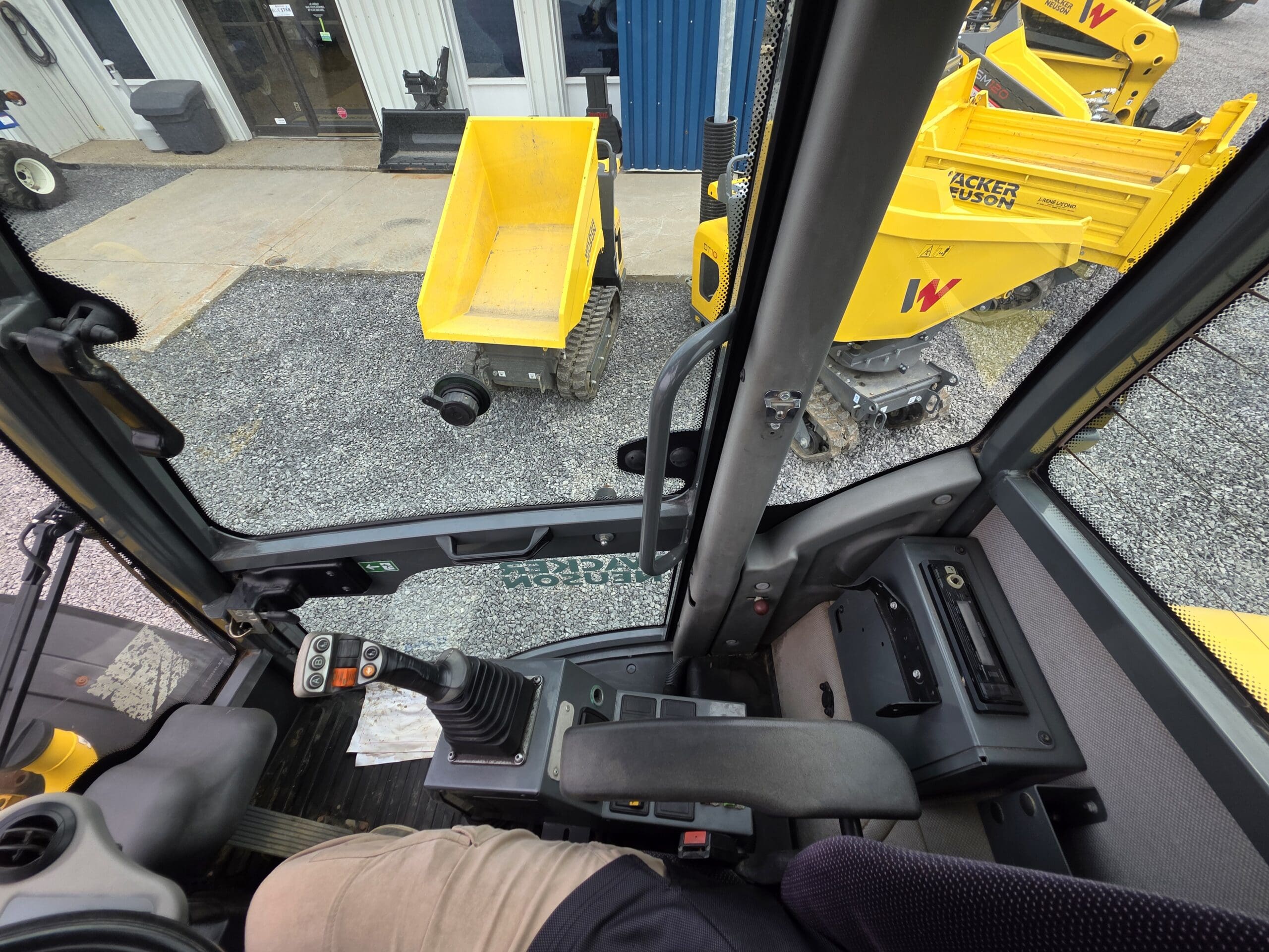 Vue de l'intérieur de la cabine d'un engin de chantier, montrant le panneau de commande, la direction et le bras de l'opérateur. À l'extérieur, des engins de chantier jaunes et du gravier sont visibles près de l'entrée d'un bâtiment.