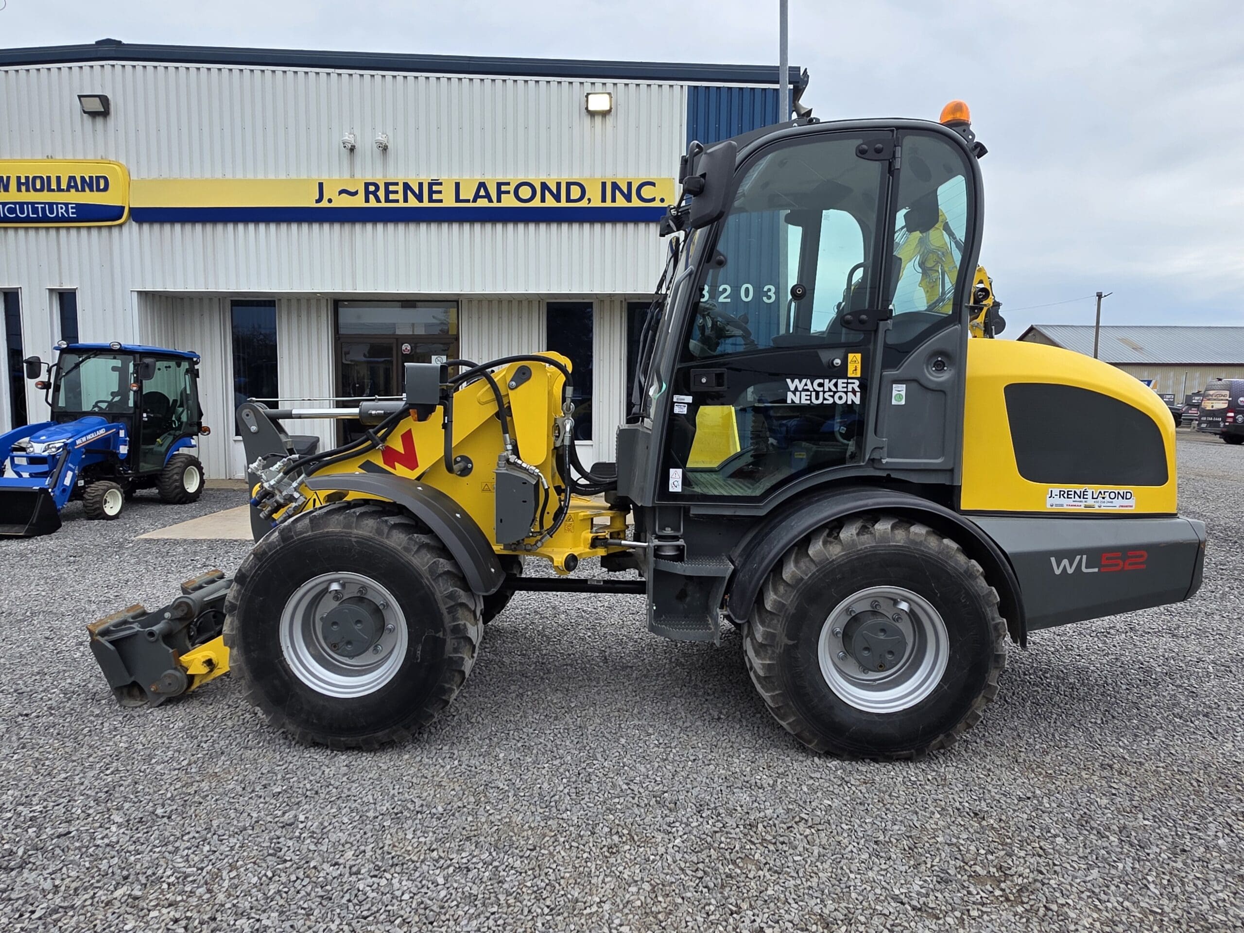 Une chargeuse sur pneus Wacker Neuson WL52 jaune et noire est garée sur du gravier devant un bâtiment portant l'inscription « J. Rene Lafond, Inc. » par temps nuageux. Un autre tracteur bleu est visible à gauche.