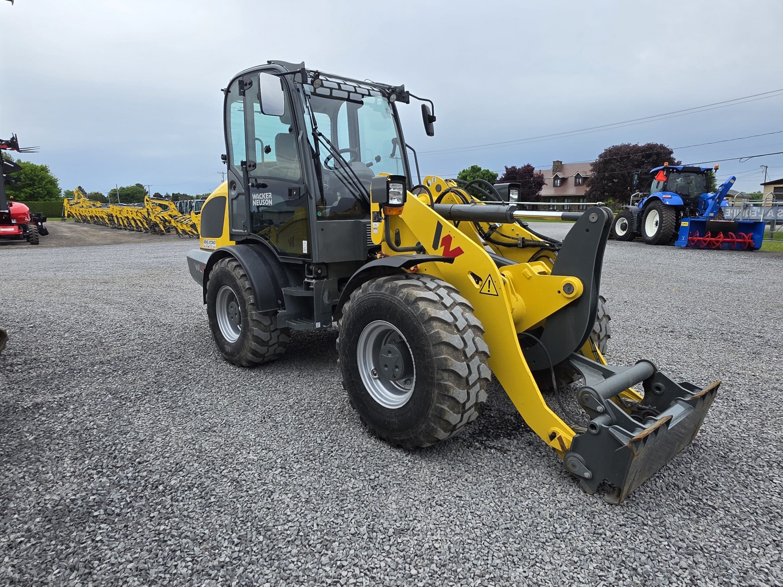 Une chargeuse sur pneus jaune et grise est garée sur un terrain en gravier, avec d'autres machines et équipements agricoles visibles en arrière-plan sous un ciel nuageux.