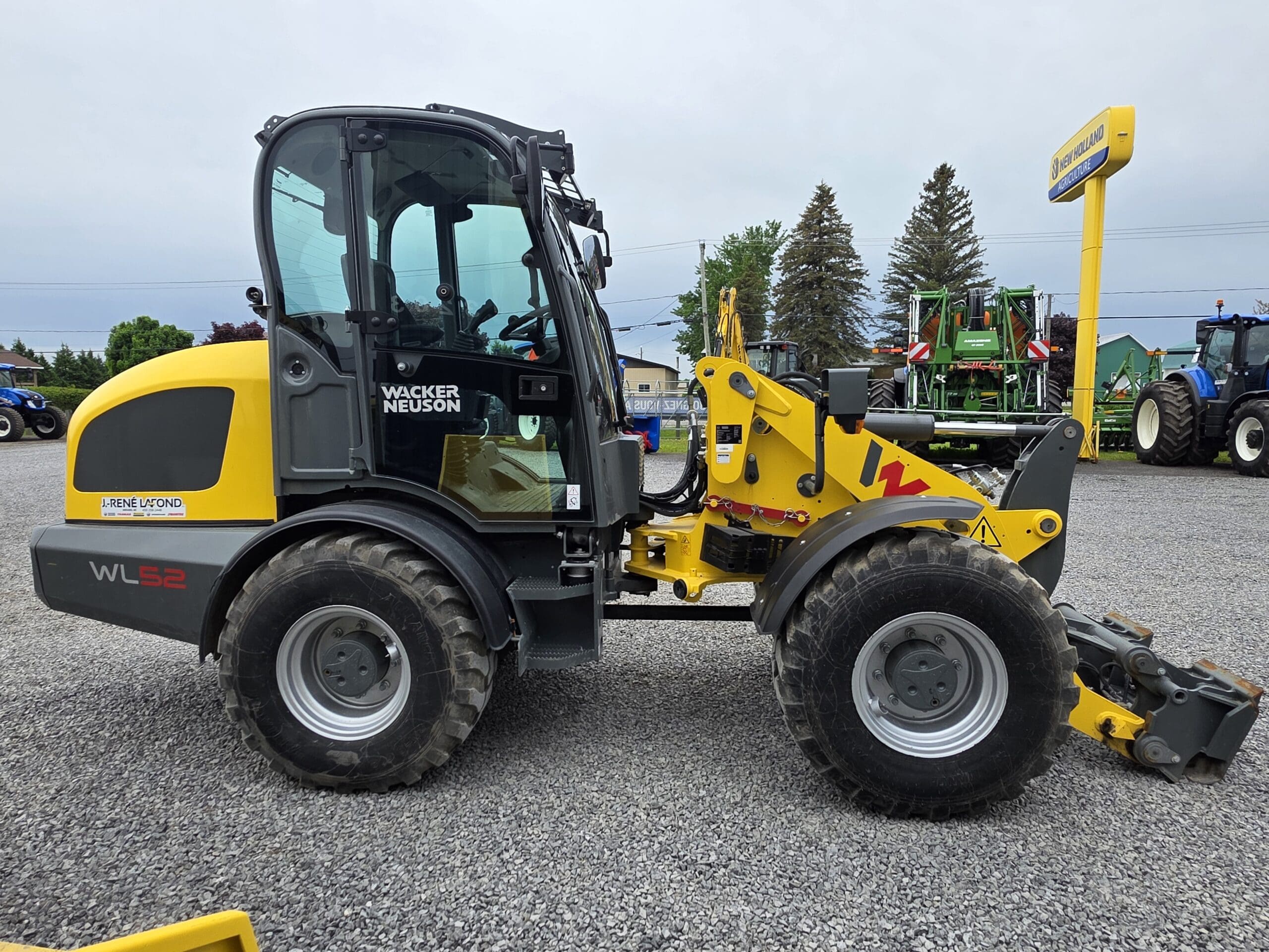 Une chargeuse sur pneus Wacker Neuson WL52 jaune et noire est garée sur du gravier, avec du matériel agricole et des arbres visibles en arrière-plan sous un ciel nuageux.