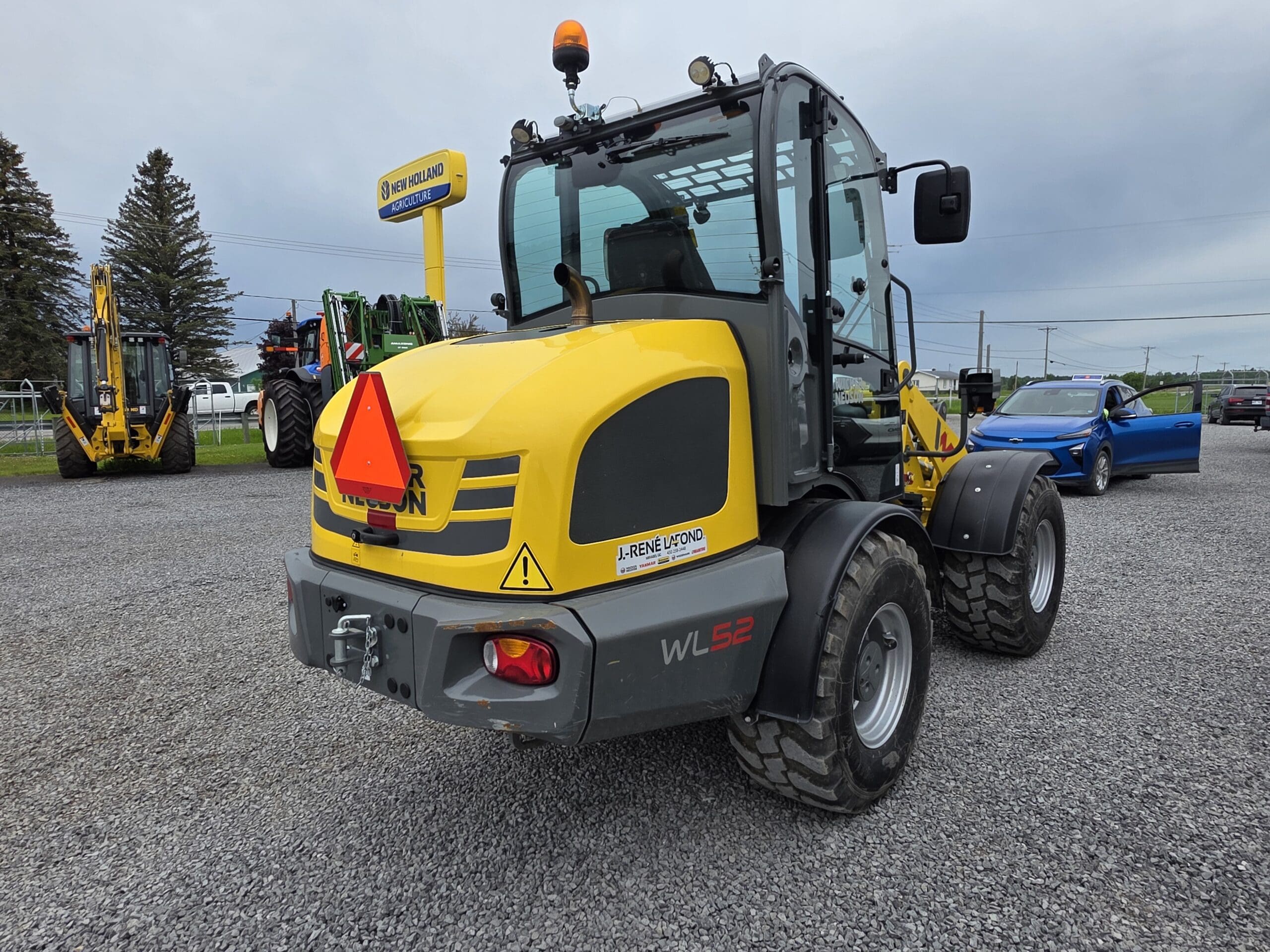 Une chargeuse sur pneus jaune Wacker Neuson WL52 avec un triangle réfléchissant rouge est garée sur du gravier, avec des tracteurs, une voiture bleue et des arbres visibles en arrière-plan sous un ciel nuageux.