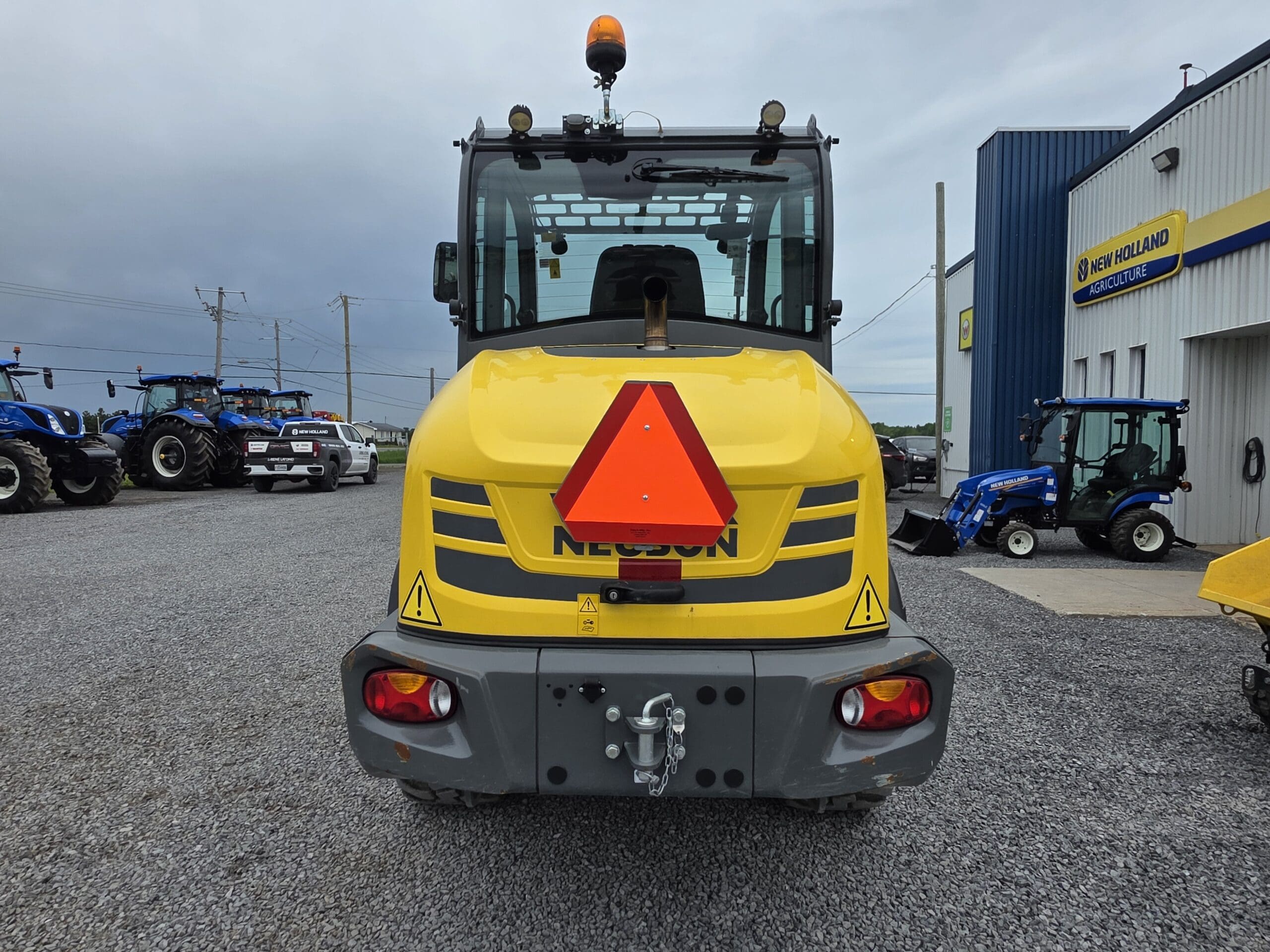 Vue arrière d'un véhicule de chantier jaune, orné d'un grand triangle réfléchissant rouge, garé sur du gravier devant une concession New Holland. D'autres tracteurs et équipements bleus sont visibles en arrière-plan.