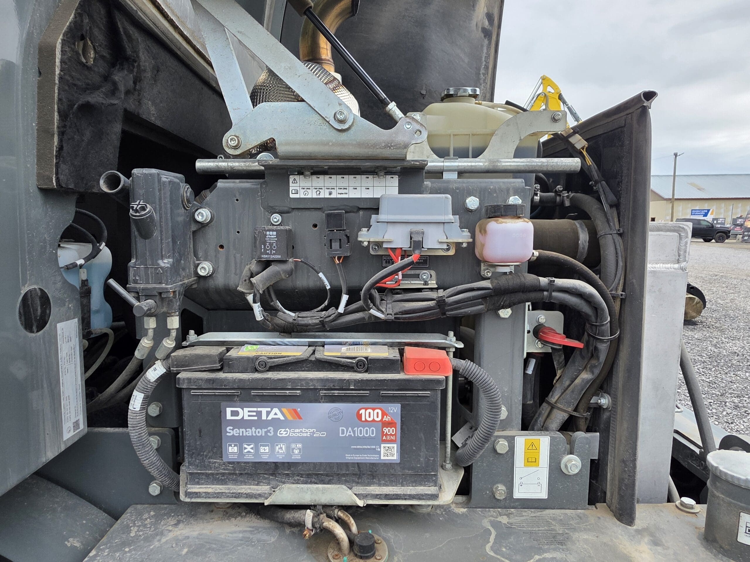 Gros plan du compartiment moteur d'un véhicule montrant le câblage, une batterie de voiture DETA, des fusibles et divers composants électroniques, avec le capot ouvert et une partie d'un bâtiment visible en arrière-plan.